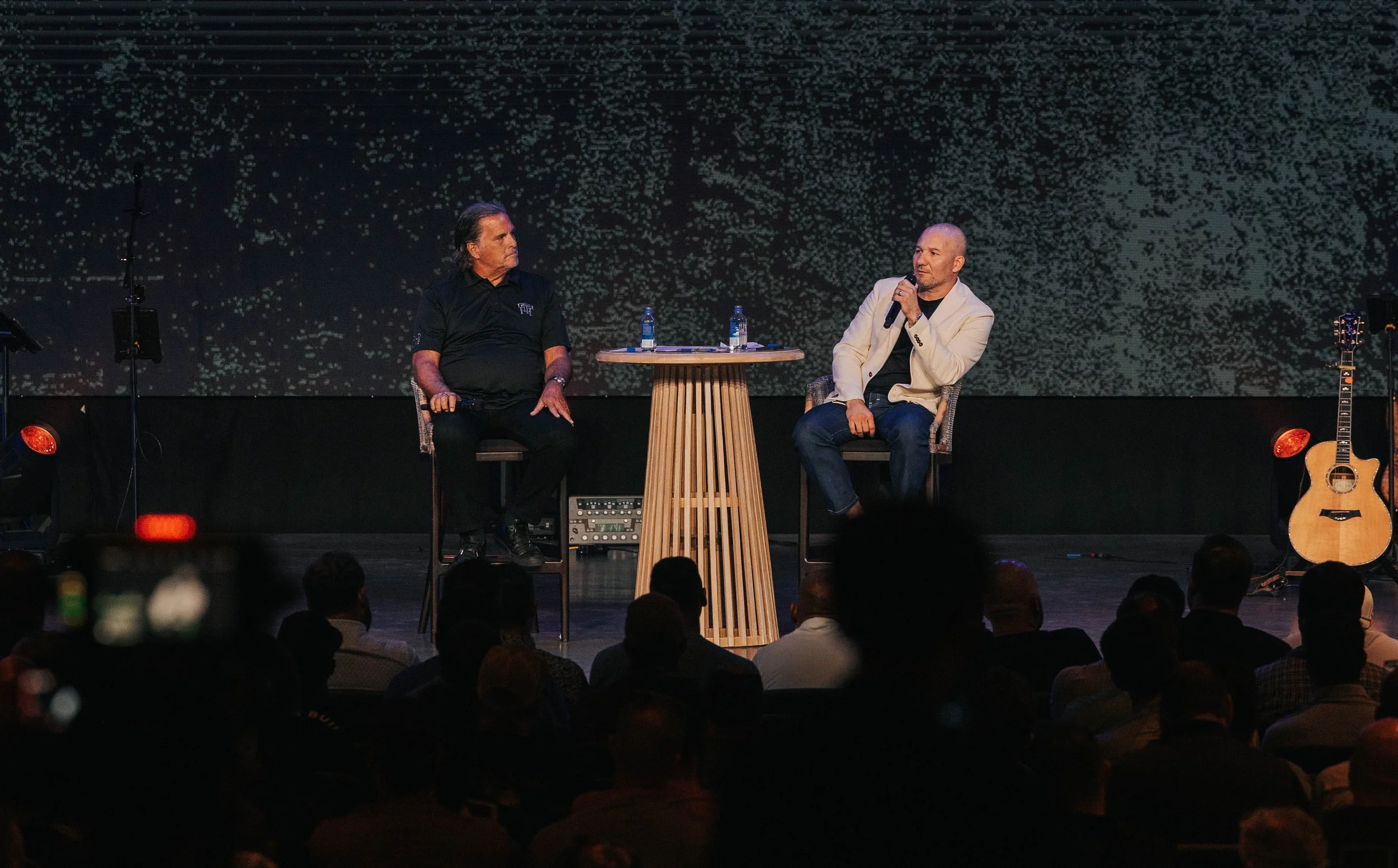 Two men on stage seated in chairs, one holding a microphone, with a small round table between them, a guitar on the right, and an audience watching, in a dimly lit auditorium.