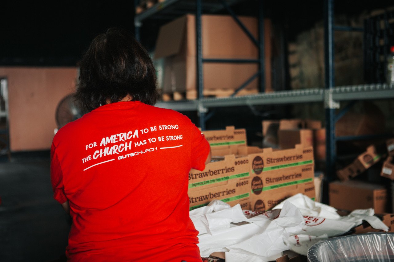 Person wearing a red shirt with white text on the back, standing in a storage or warehouse area with shelves filled with cardboard boxes of strawberries.