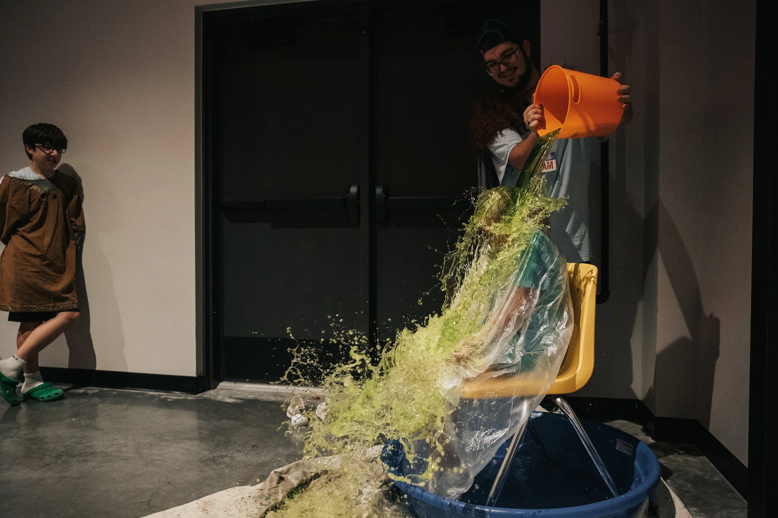 Person pouring a bucket of yellow and green slime into a plastic bag, causing the slime to spill out and splash onto the floor, with a woman standing nearby watching.