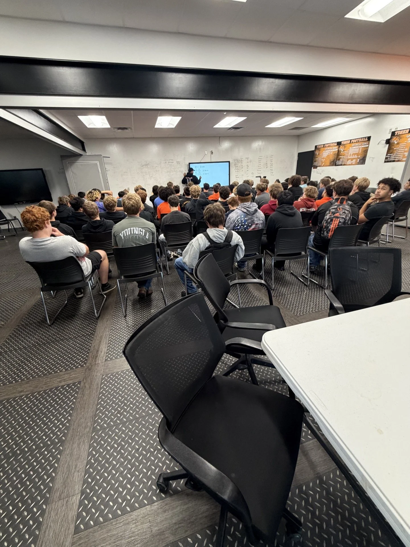 Classroom filled with students listening to a teacher at a whiteboard with notes. The room has black chairs and white walls with posters.