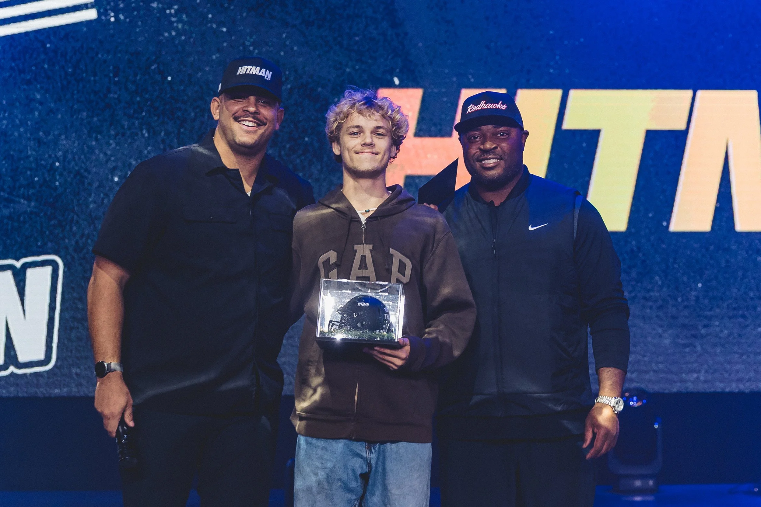 Three men standing on a stage, with one wearing a hoodie and holding an award, flanked by two men in athletic jackets and caps, smiling for the photo.