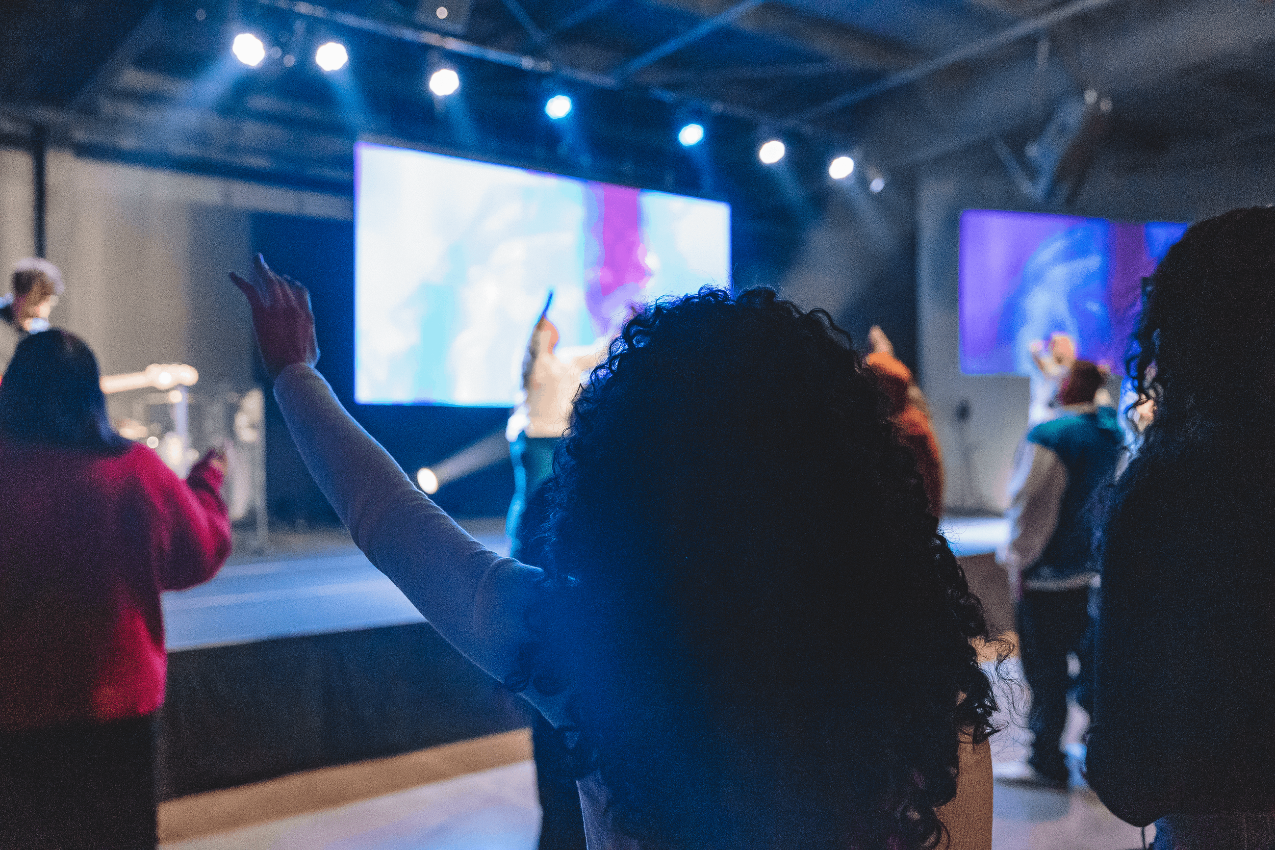 A speaker on stage speaking to an audience in a darkened venue, with some audience members standing and others seated.
