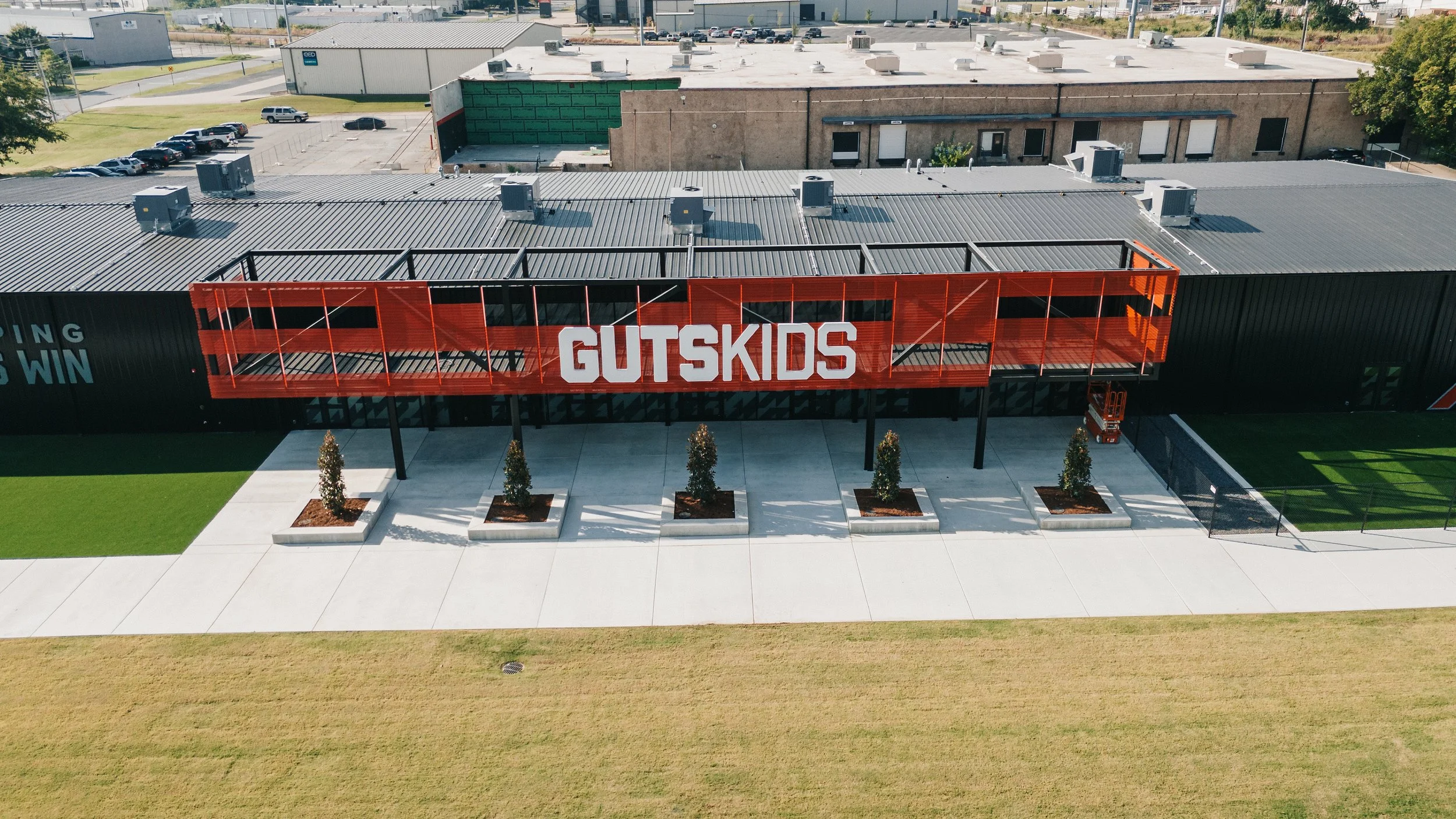 Aerial view of a modern building with a large sign reading 'GUTSKIDS' on a red metal structure across the front. The building has a black exterior with a gray metal roof, and there are five young trees planted in concrete planters in front of it. To the right, there is a small fenced area with artificial turf, and to the left, a sidewalk with a grassy strip. Parking lots and other industrial buildings are visible in the background.
