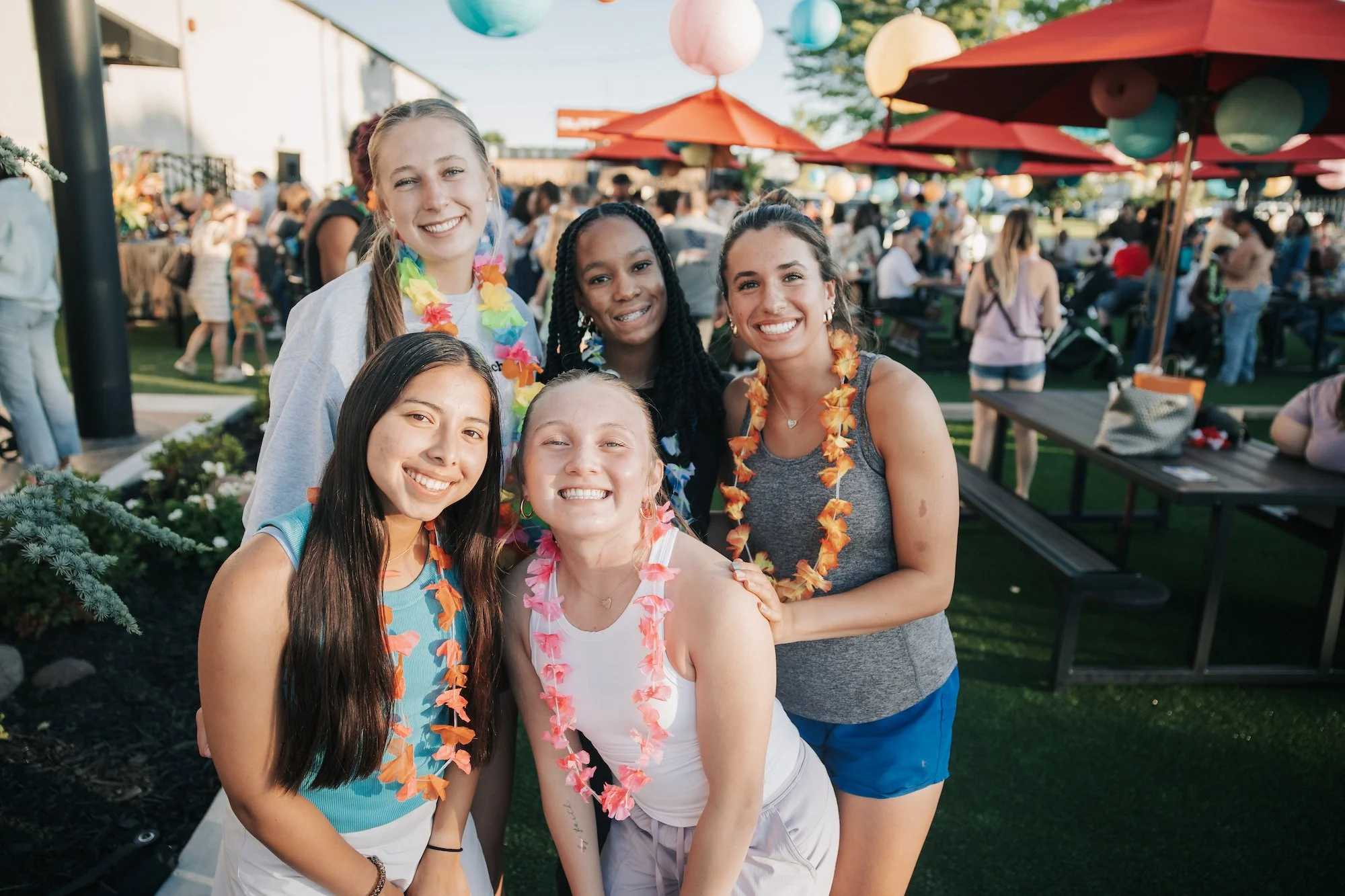 A group of five young women smiling and wearing colorful flower leis at an outdoor gathering with picnic tables and umbrellas in the background.