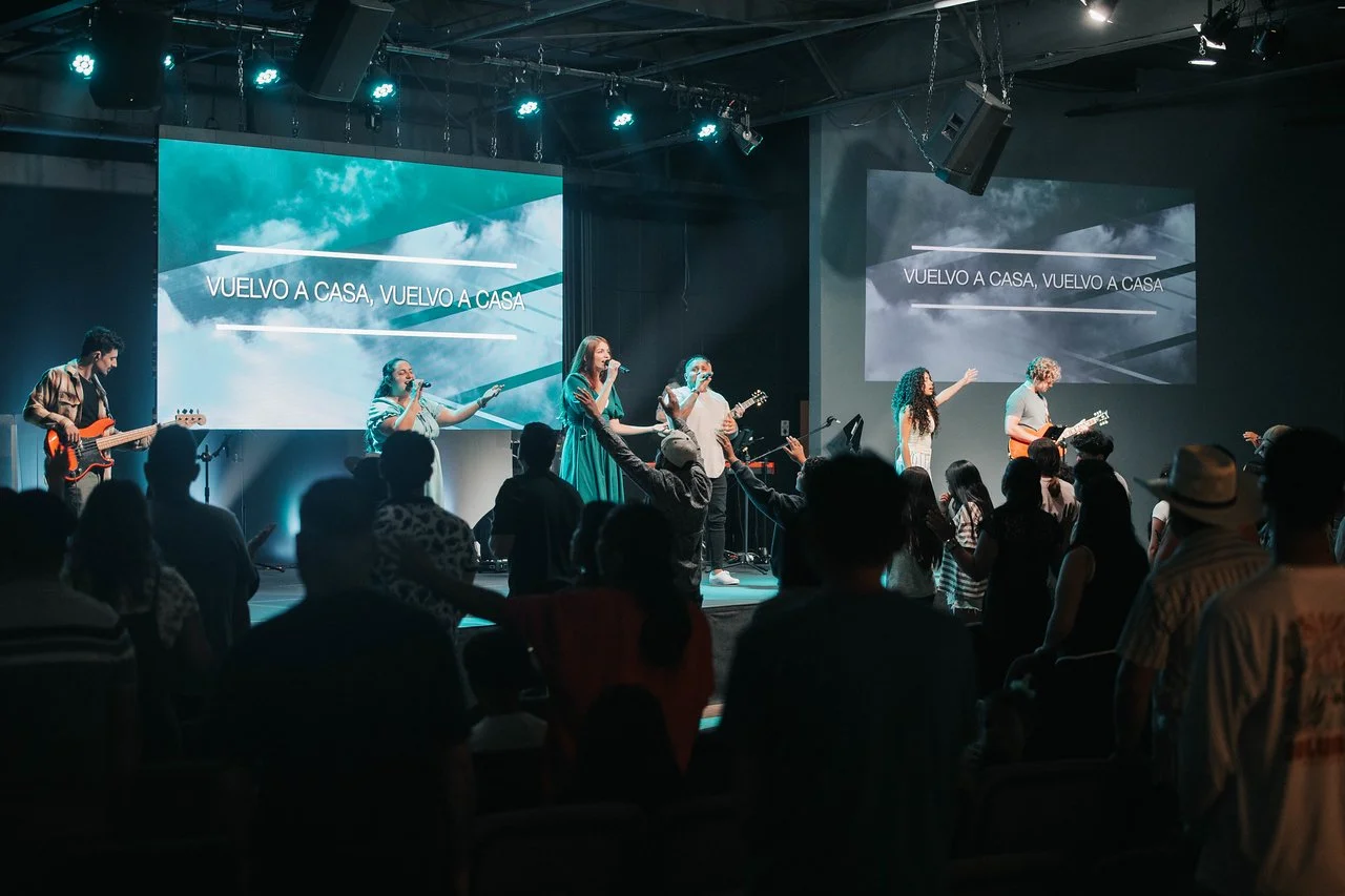 A band performs on stage with five musicians, including a guitarist, two singers, a bassist, and a drummer, during a live concert. The audience is standing and watching, some with hands raised. Large screens behind the band display the Spanish phrase 'VUELVO A CASA, VUELVO A CASA' against a sky and cloud background.