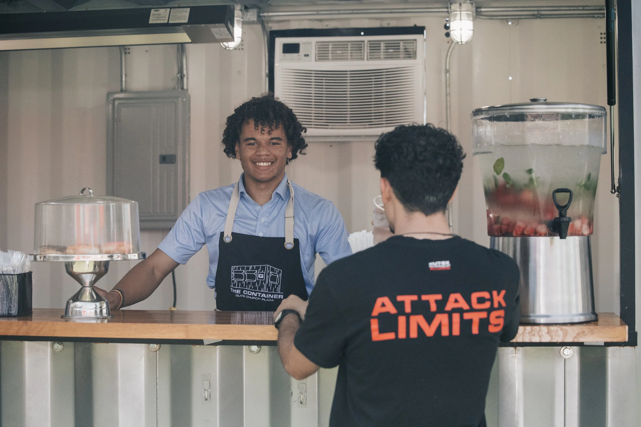 A young man with curly dark hair smiling behind a counter, wearing a light blue shirt and an apron, talking to a customer who has short dark hair, wearing a black shirt with red text that says 'Attack Limits,' in a food stand or cafe setting with a container of water with fruit and herbs on the right side.