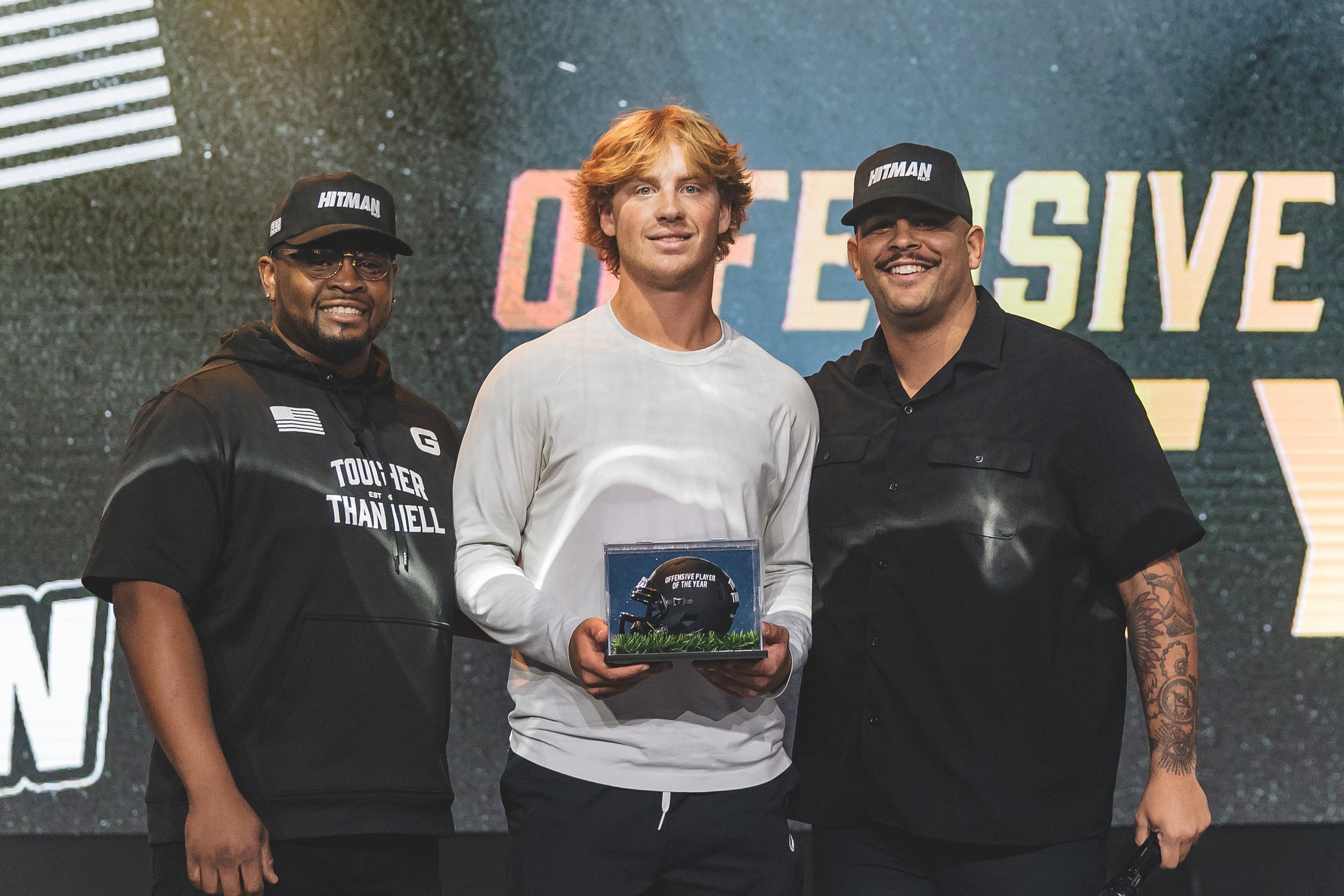 Three men smiling and standing together; one in the middle holding an award, at a sports event, with a backdrop displaying the word "OFFENSIVE".