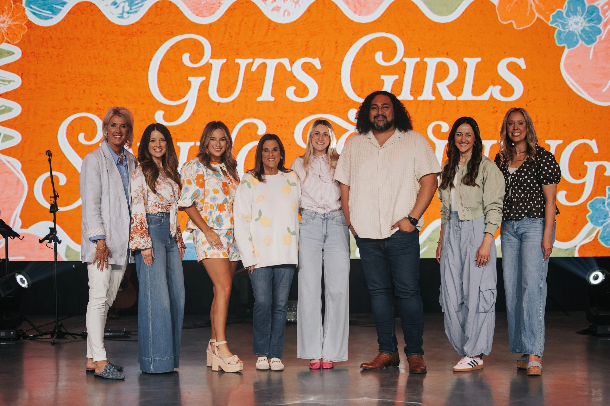 Group of nine women and one man standing on stage with a bright orange background that says "Guts Girls Succeeding" in large white letters, wearing casual and colorful clothing.