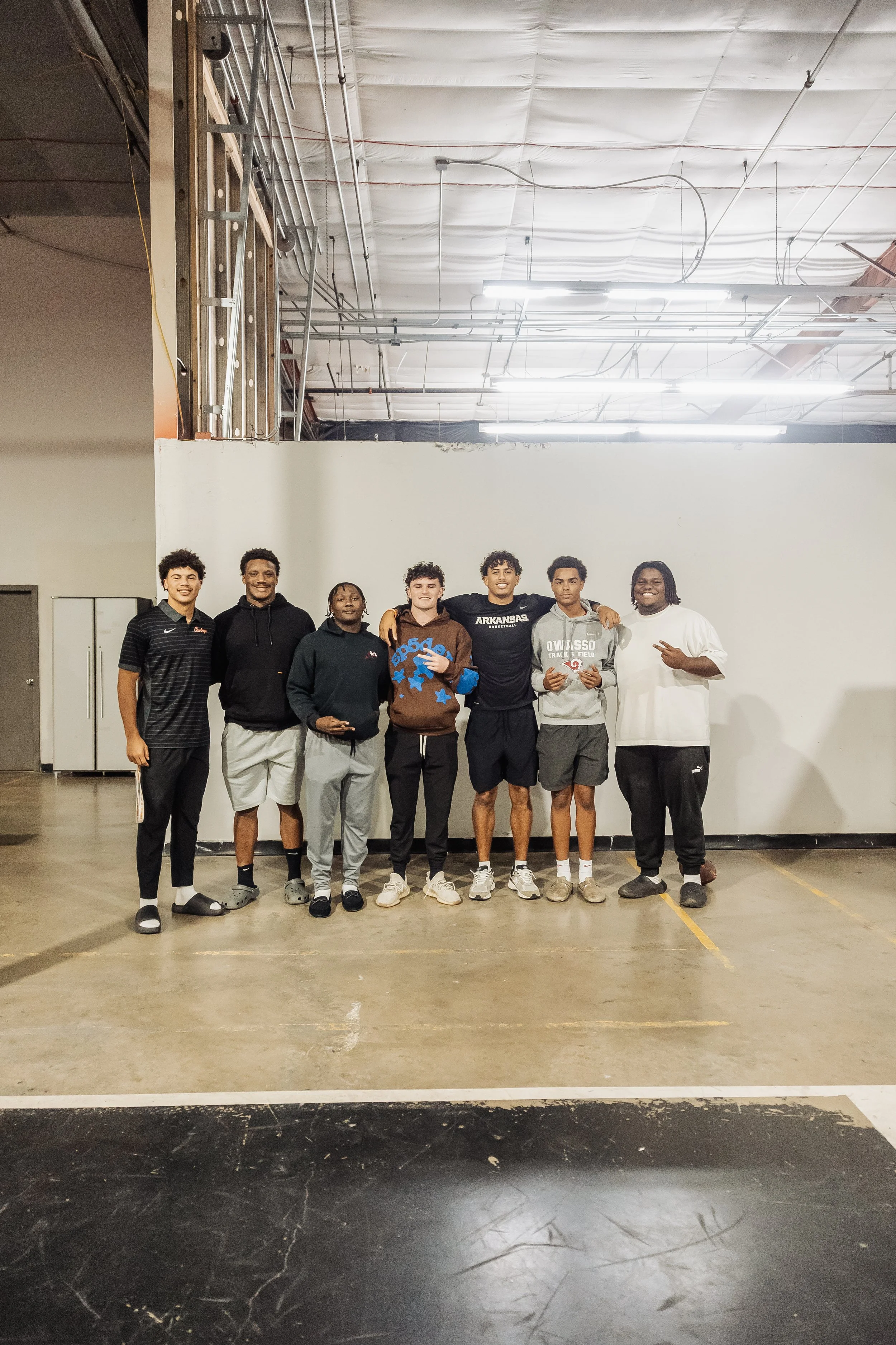 A group of nine young men standing together in a gymnasium, posing for a photo. They are dressed in sporty casual clothing, with some in hoodies, shorts, and sneakers. The background shows a white wall and a ceiling with exposed electrical wires and 