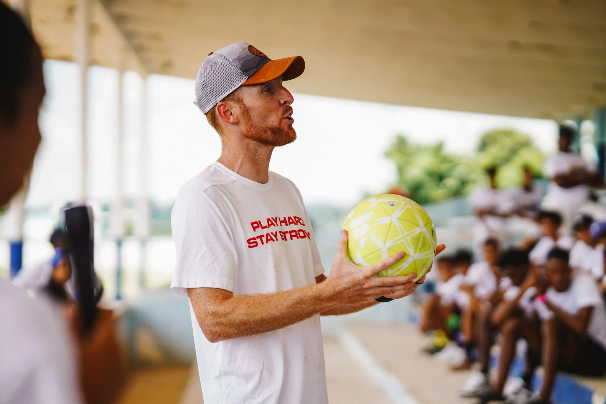 A man with red hair and beard, wearing a gray and orange baseball cap and a white t-shirt, is holding a yellow and green volleyball and appears to be speaking. In the background, a group of children in white shirts are sitting on bleachers at an indo