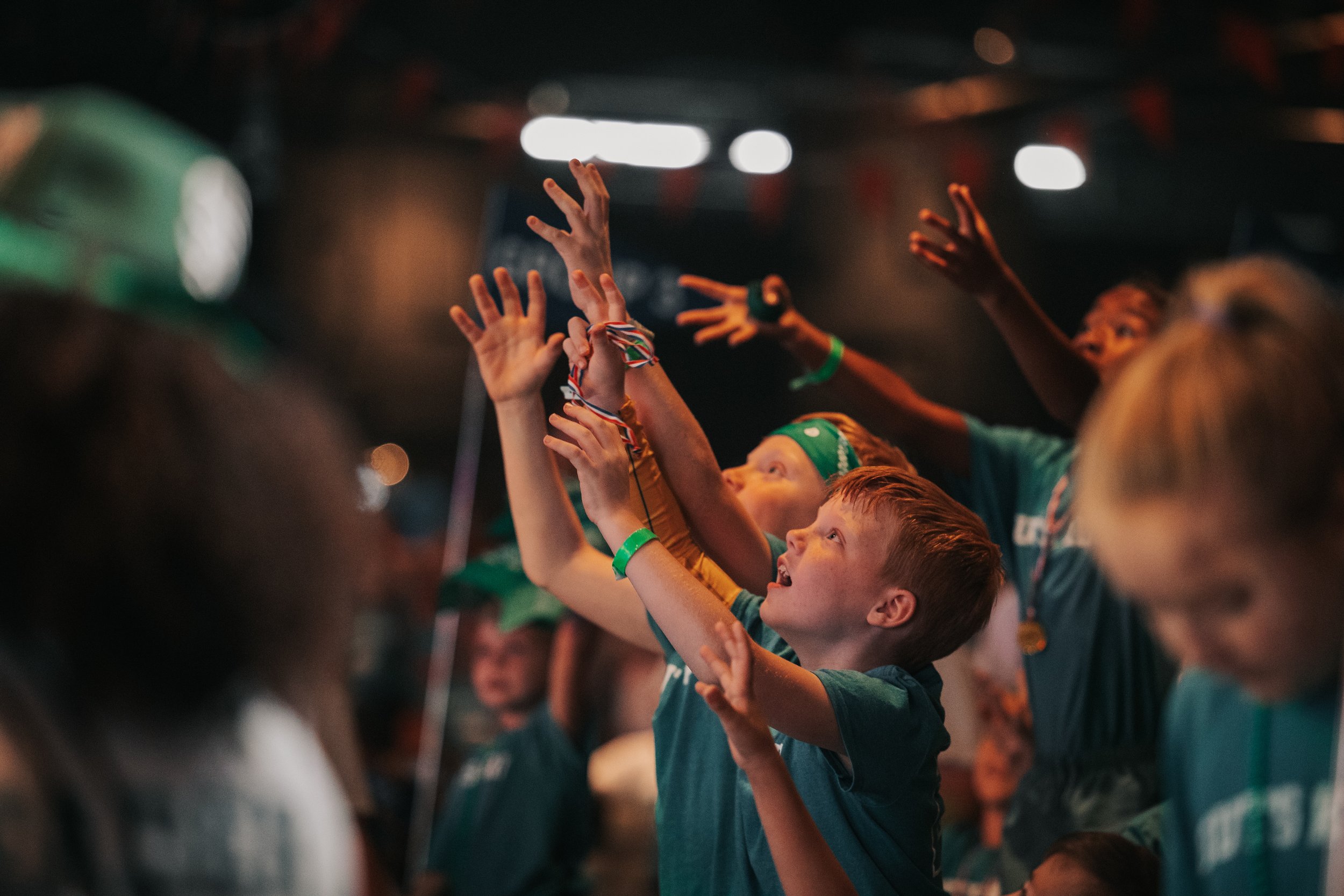 Children at an indoor event wearing orange shirts with 'GUTS KIDS' printed on them, some waving green flags and others holding green star-shaped balloons, with green and yellow bunting hanging from the ceiling.
