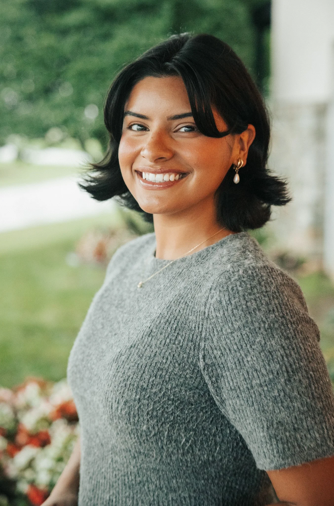 A woman with long dark hair smiling, wearing a white embroidered blouse, standing outdoors with a blurred background of buildings and plants.