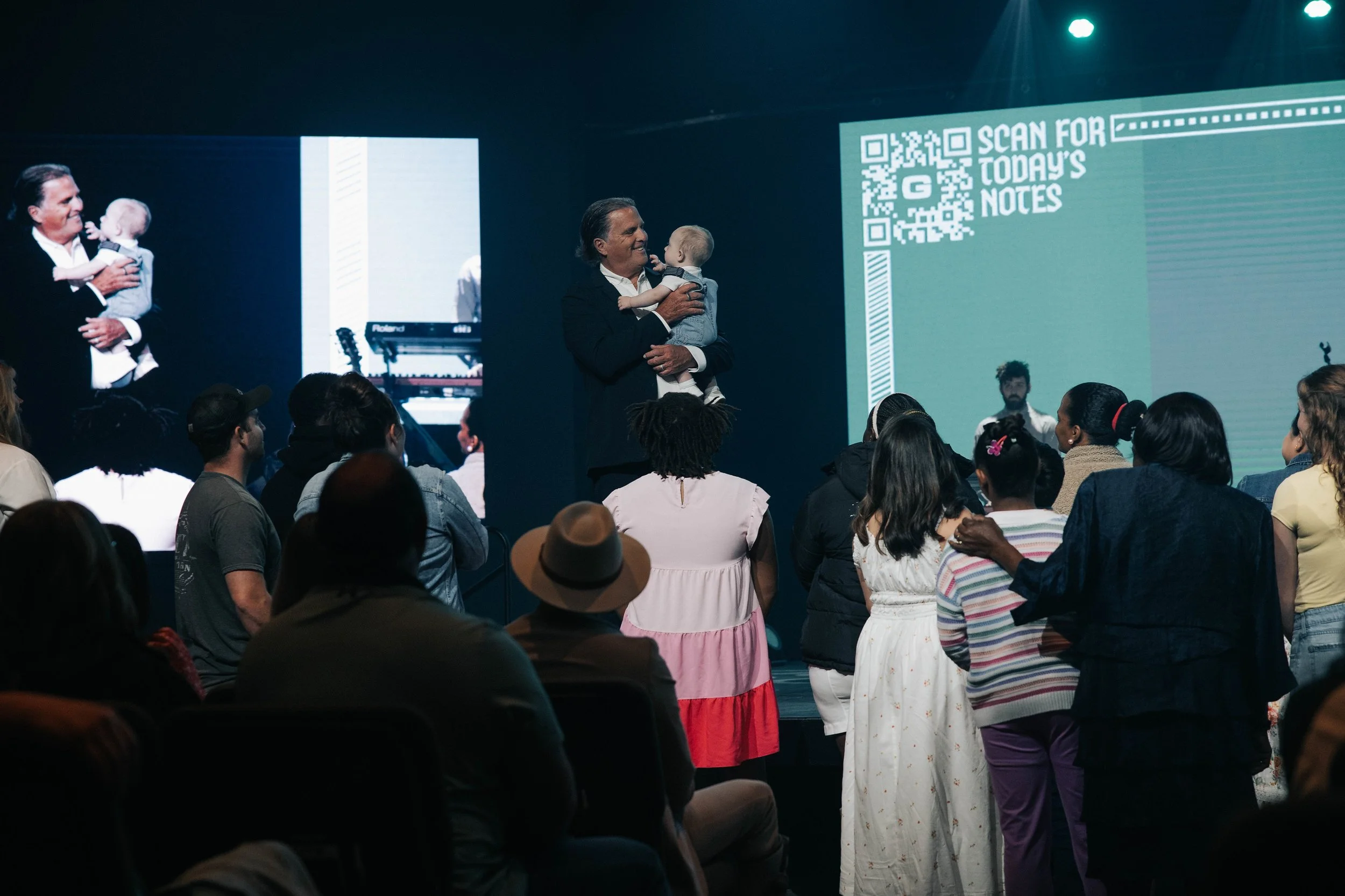 Woman holds young child on stage during event with audience and large screen showing a QR code and text in the background.