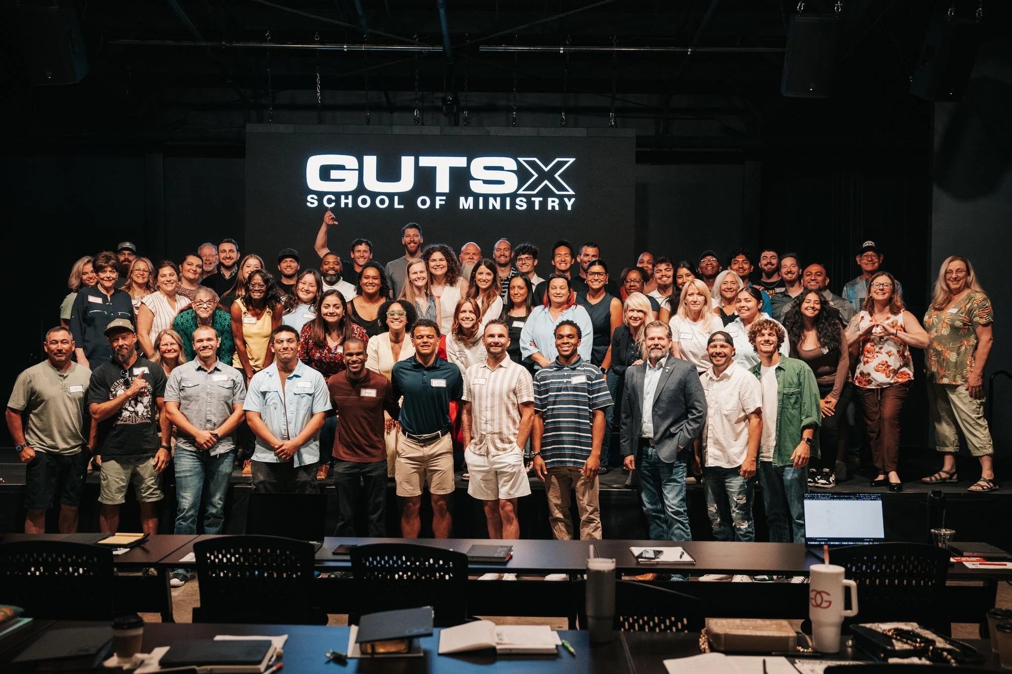 Large group of people on stage at GUTS X School of Ministry event, smiling and posing for photo, with black backdrop and digital screen displaying the event name.