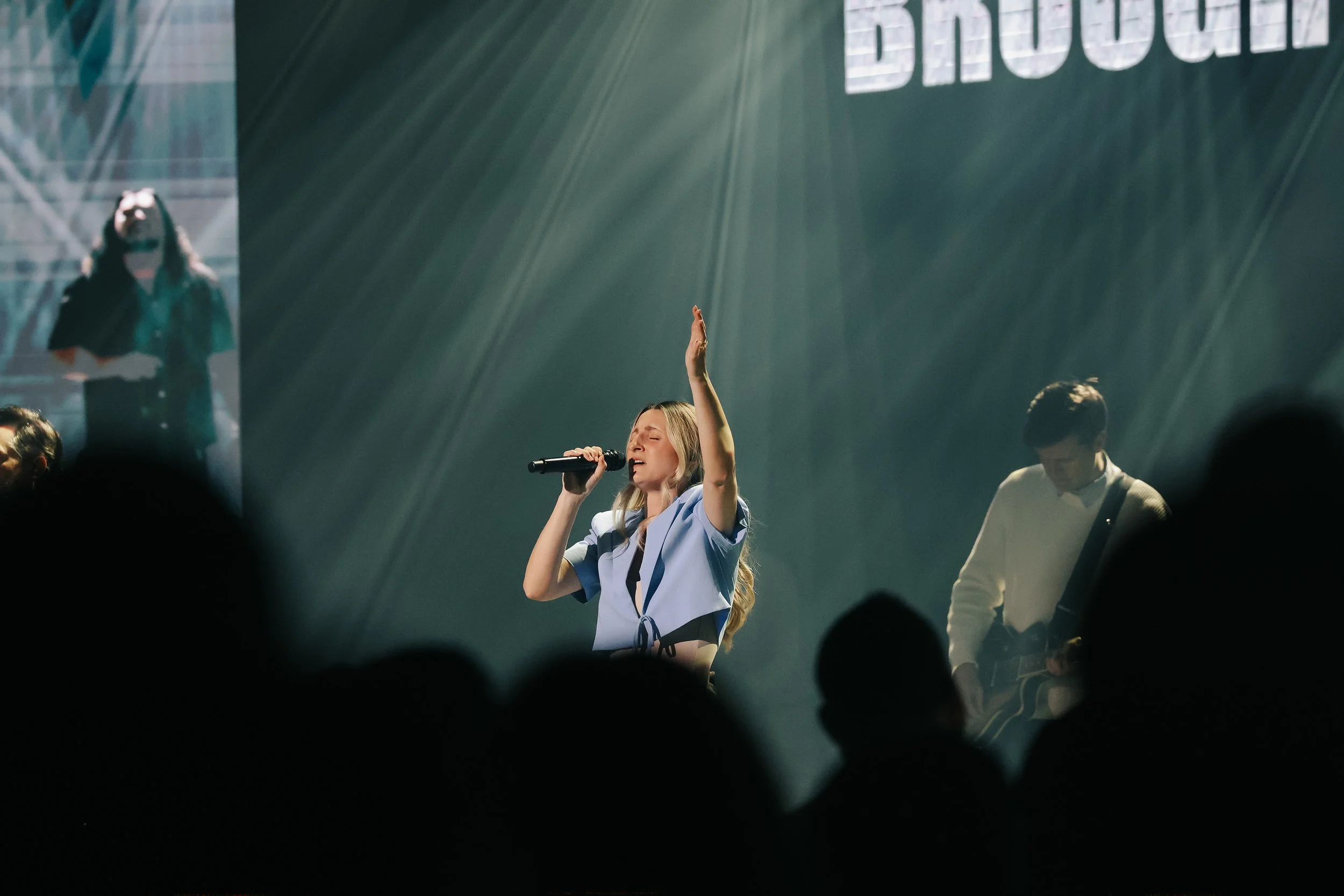 A woman singing into a microphone on stage with her eyes closed and one arm raised, while a man plays guitar beside her. There is a large screen behind them with partially visible text.