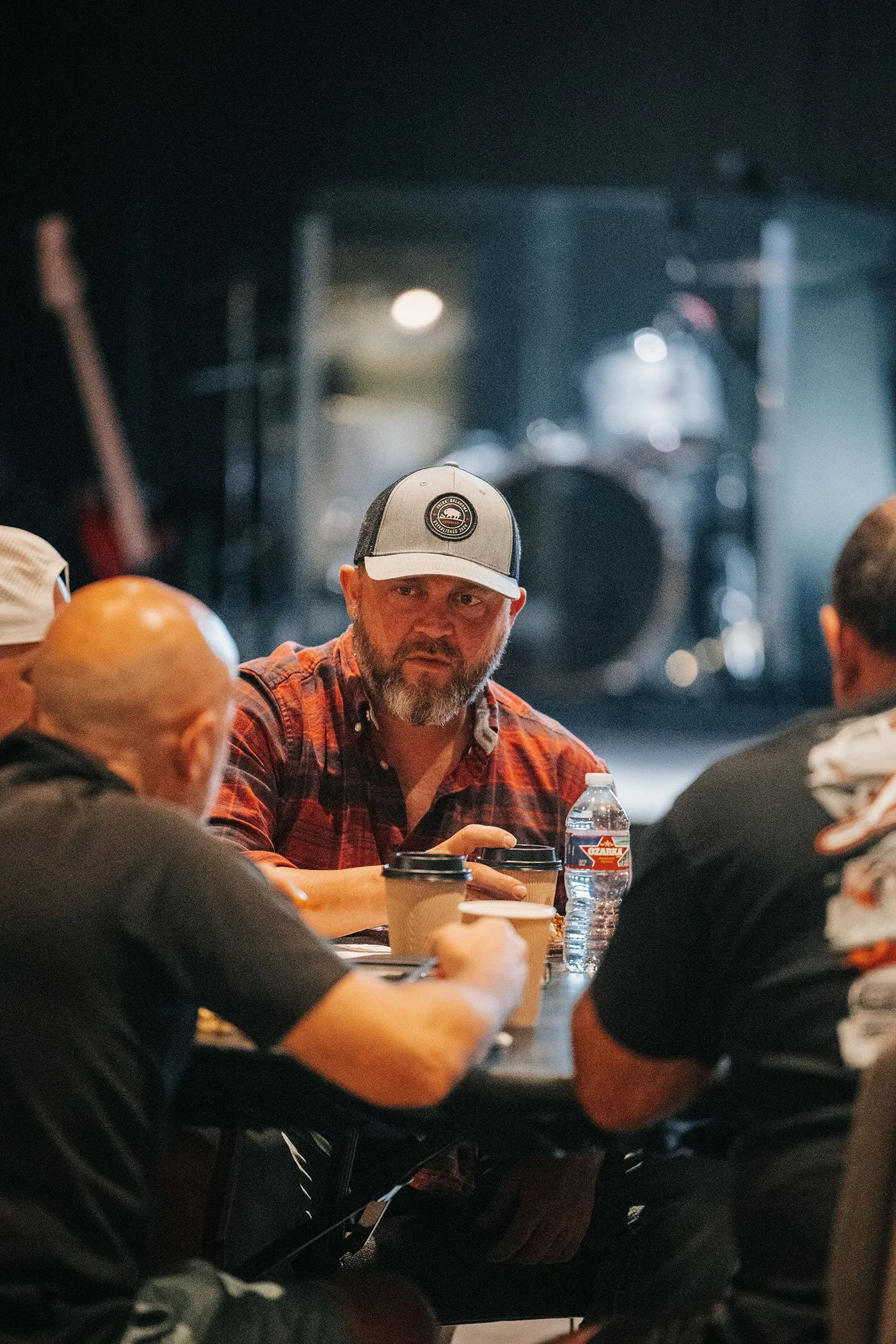 Man with beard wearing a gray and black baseball cap and red plaid shirt, sitting at a table with other people, in a venue with musical instruments in the background.