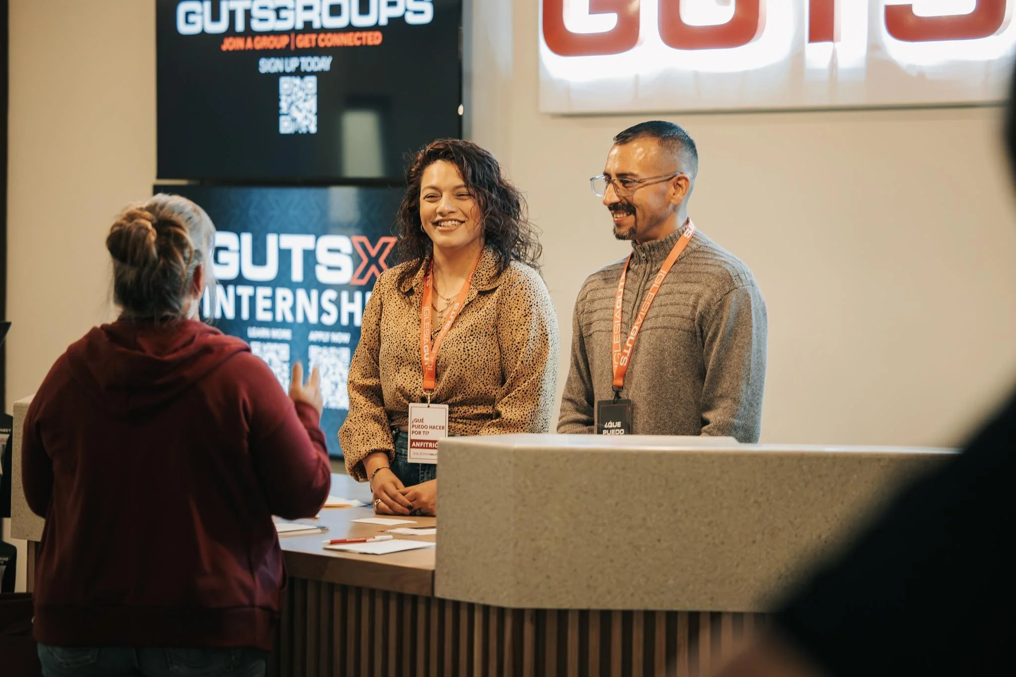 A woman with gray hair in a red jacket is talking with two people, a woman with and curly hair in a beige shirt and a man with glasses and a tan sweater, at a conference registration desk with a sign behind them that reads 'GUTS X INTERNSHIP.'