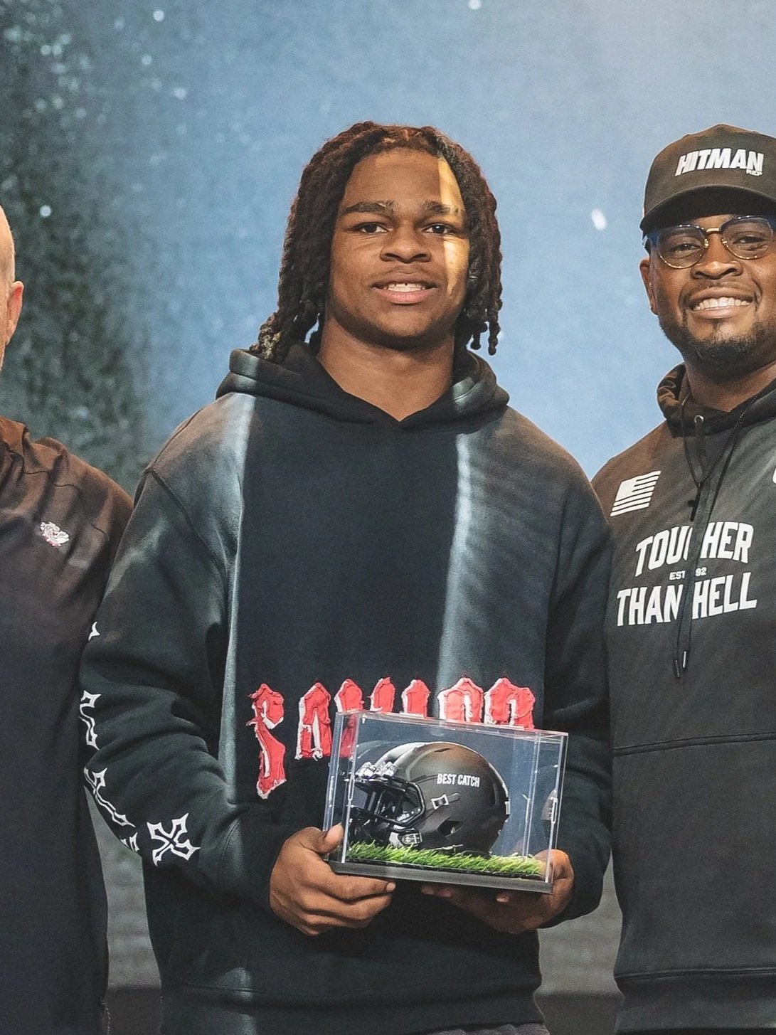 Young man with dreadlocks holds a mini football helmet display case that says 'Best Catch.' He is wearing a black hoodie with red lettering and is standing with two smiling men, one on each side, in casual sportswear.