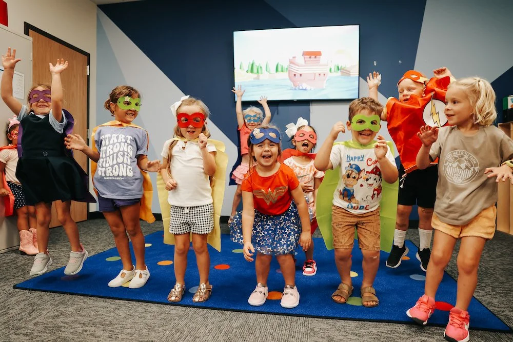 Children dressed as superheroes and wearing masks, playing and posing in a classroom or play area.