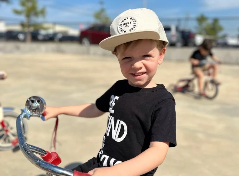 A young boy with a white cap and a black T-shirt smiling while sitting on a bicycle in a parking lot, with another child riding a bike in the background.