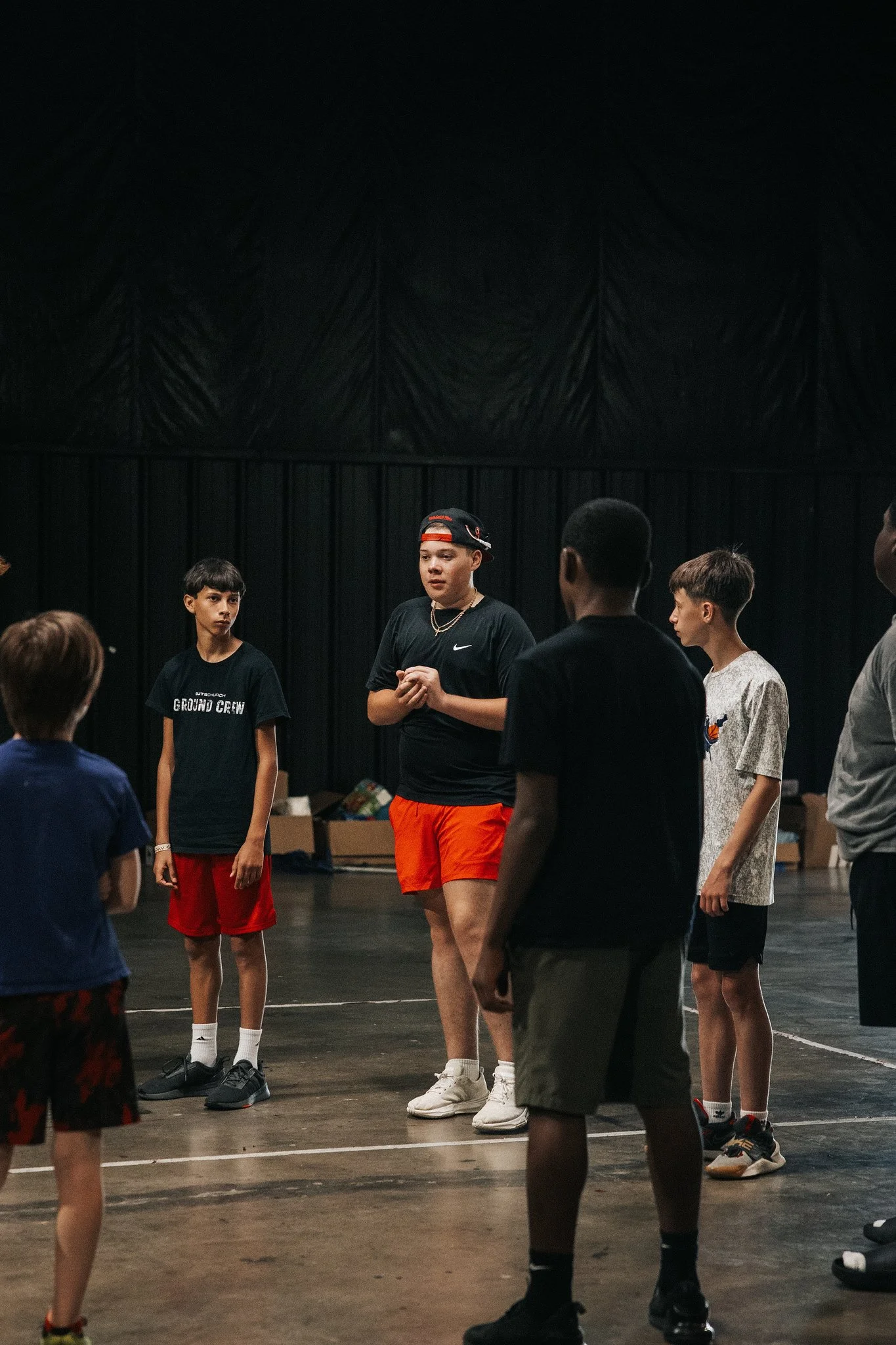 Group of young boys and a teenage girl standing in a circle on an indoor basketball court with black curtains in the background, possibly during a team meeting or practice.