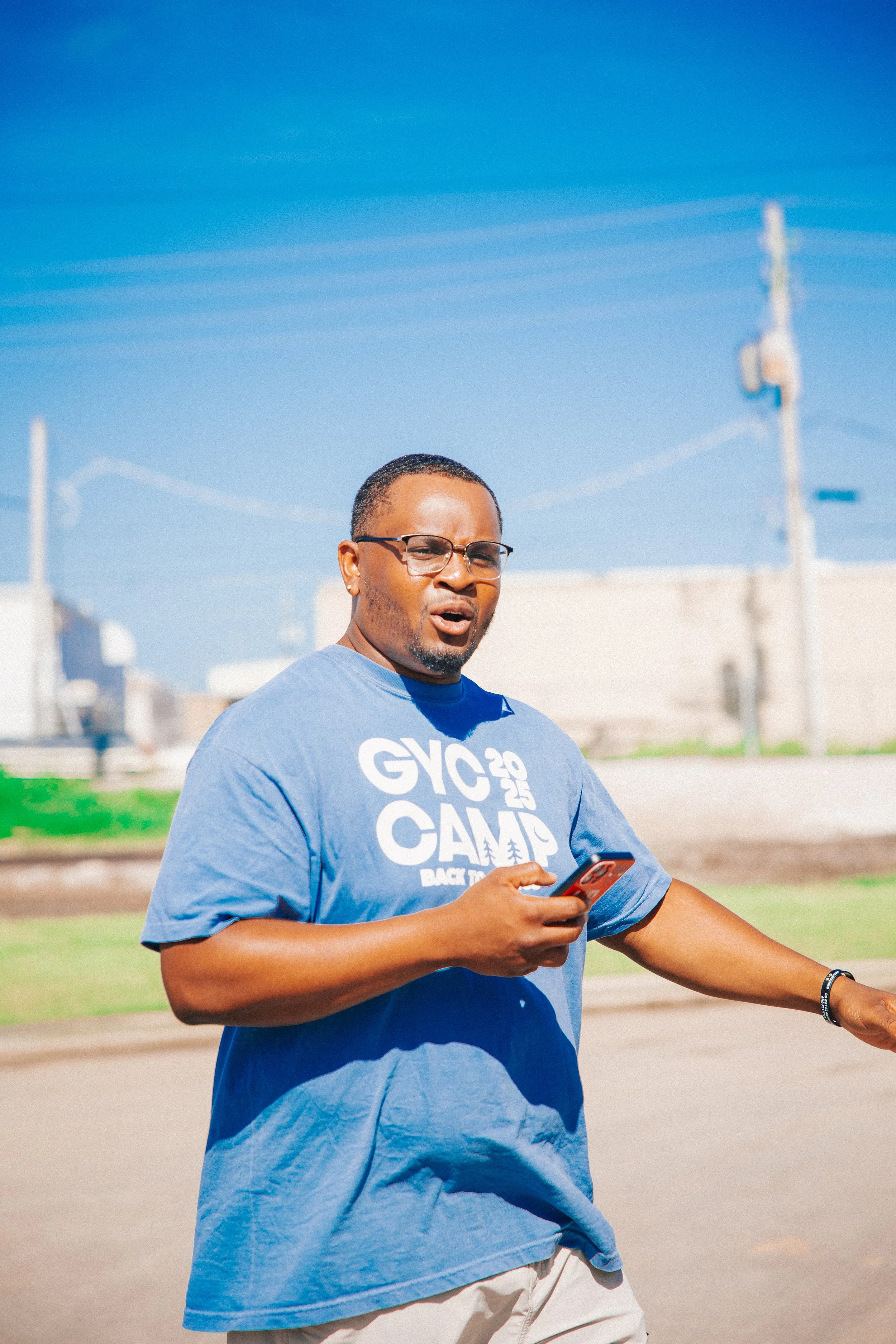 A man in glasses wearing a blue T-shirt with 'GYC 2025 CAMP BACK TO' printed on it, holding a phone, outdoors on a sunny day with power lines and a building in the background.