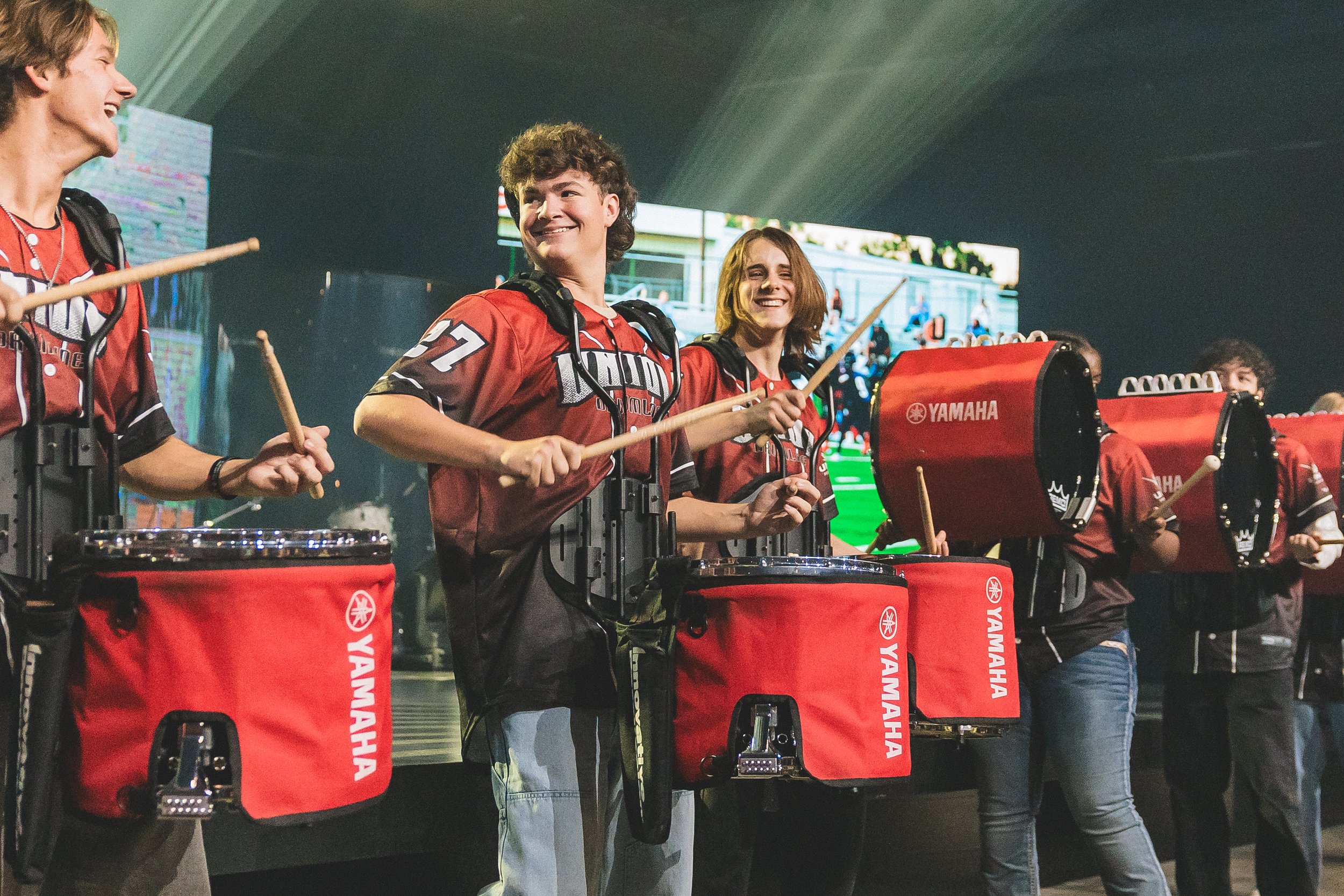 A group of young people wearing matching red and black jerseys with Yamaha branding are playing drums in a band on stage, smiling and interacting with each other.