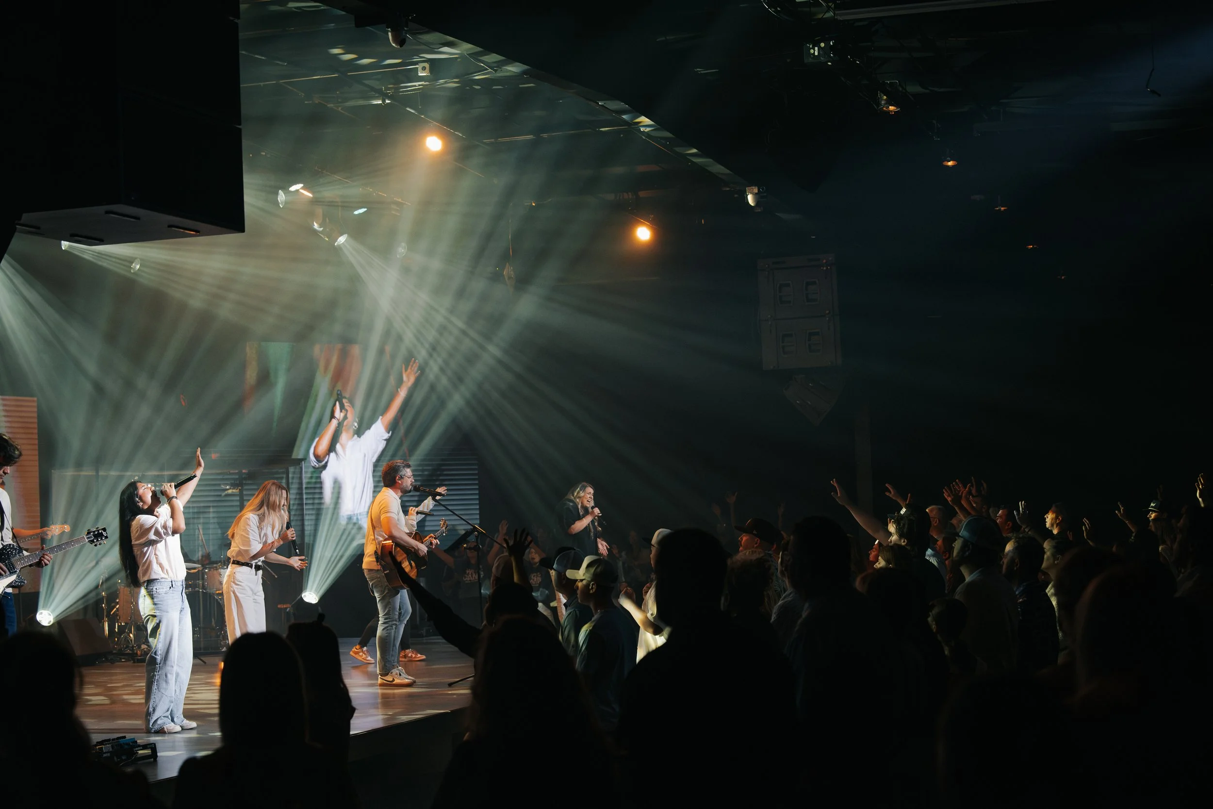 A live music performance on stage with a band, singers, and an audience in a dark venue with stage lighting and smoke effects.