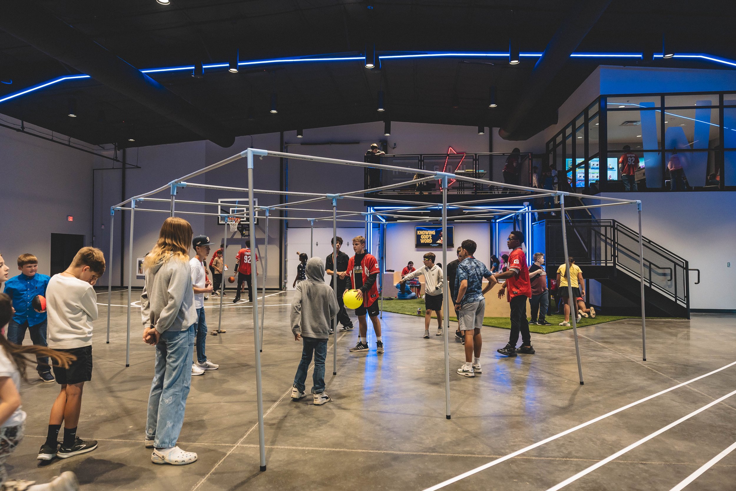 Children and teenagers playing basketball inside an indoor sports facility with a trampoline area, some perform basketball shots, others watch or wait, background includes stairs, glass balcony, and neon lights.