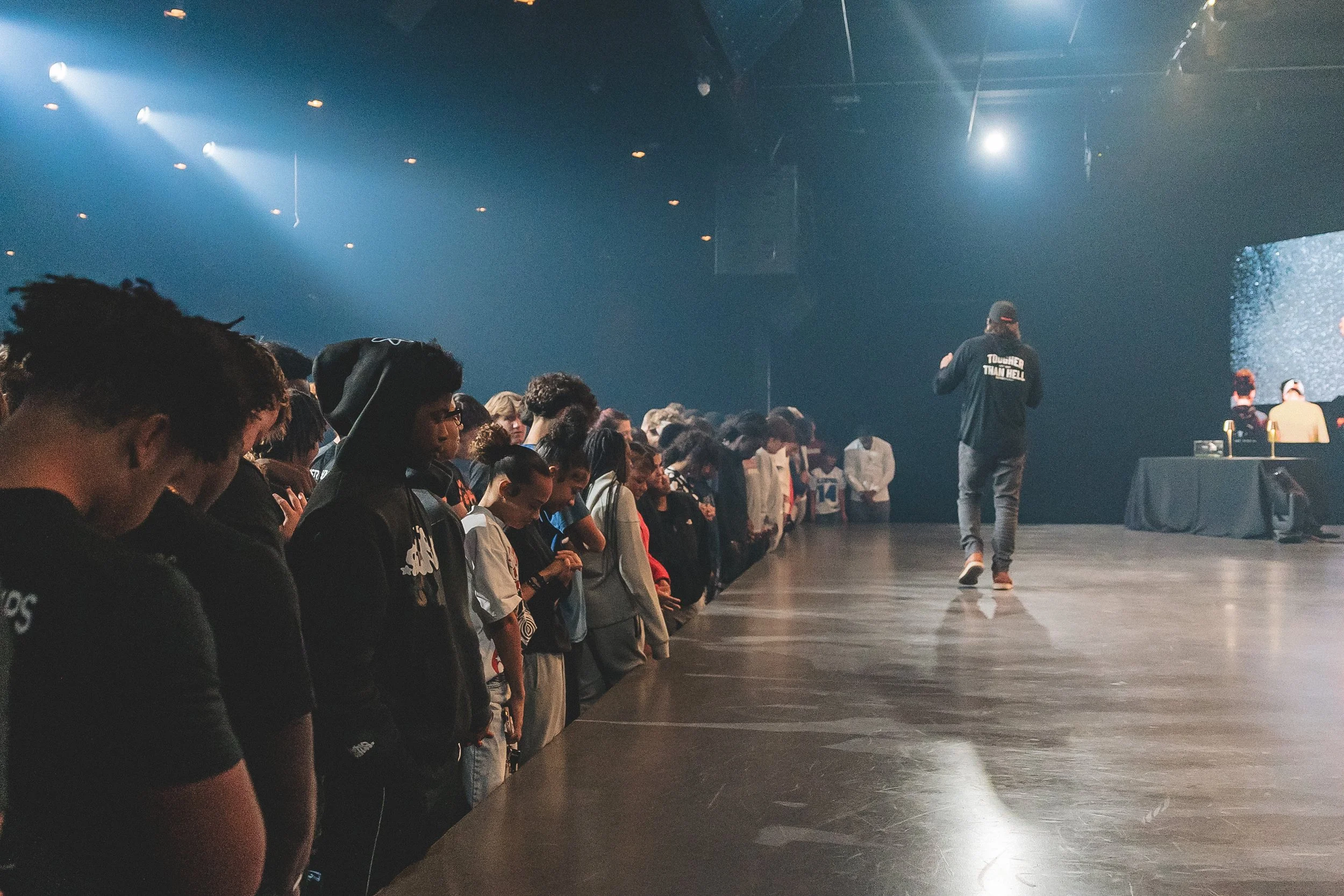 A group of people standing with heads bowed, praying or reflecting, in an indoor venue with a performer on stage facing away, wearing a hoodie that says 'Trouble Than Hell'. The stage has dark lighting with some bright spotlights, and there is a tabl