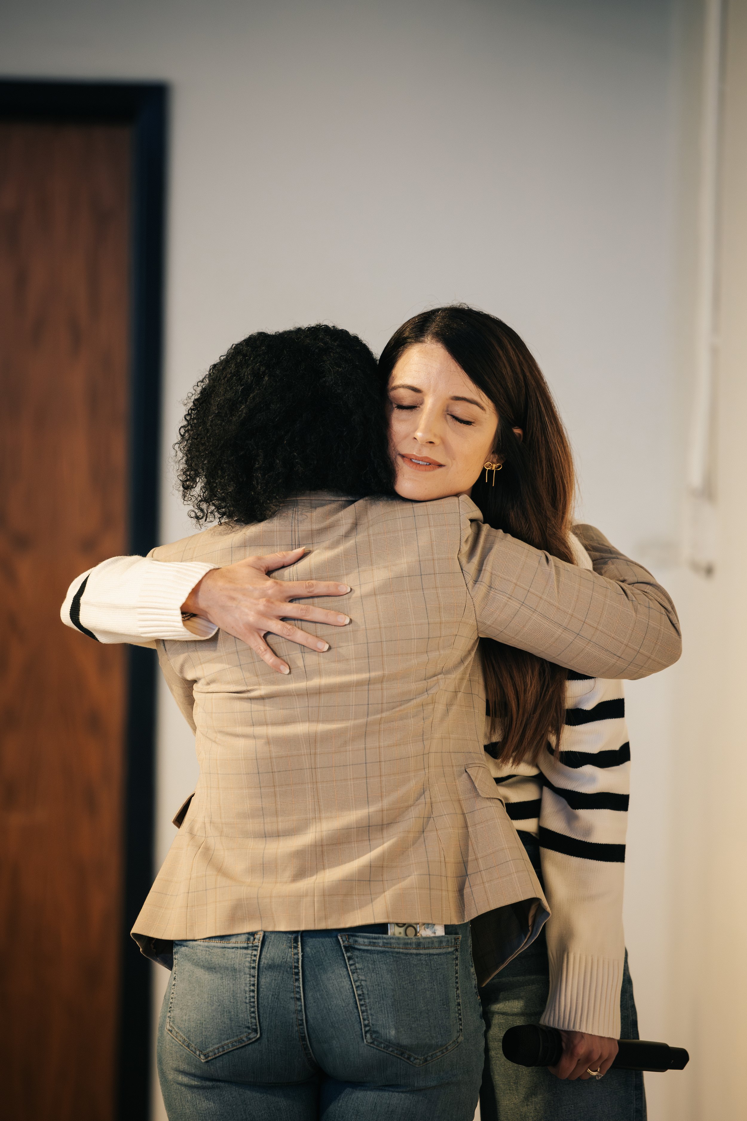 Two women hugging. One woman faces away, and the other faces toward the camera with eyes closed, holding a microphone.