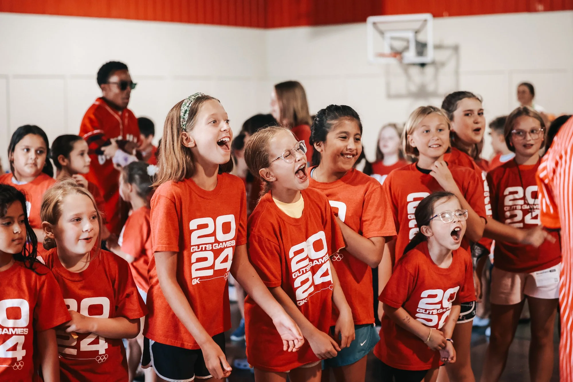 A group of young girls wearing red T-shirts with the logo of the 2024 Paris Olympics, some are smiling and laughing while standing in a gymnasium.