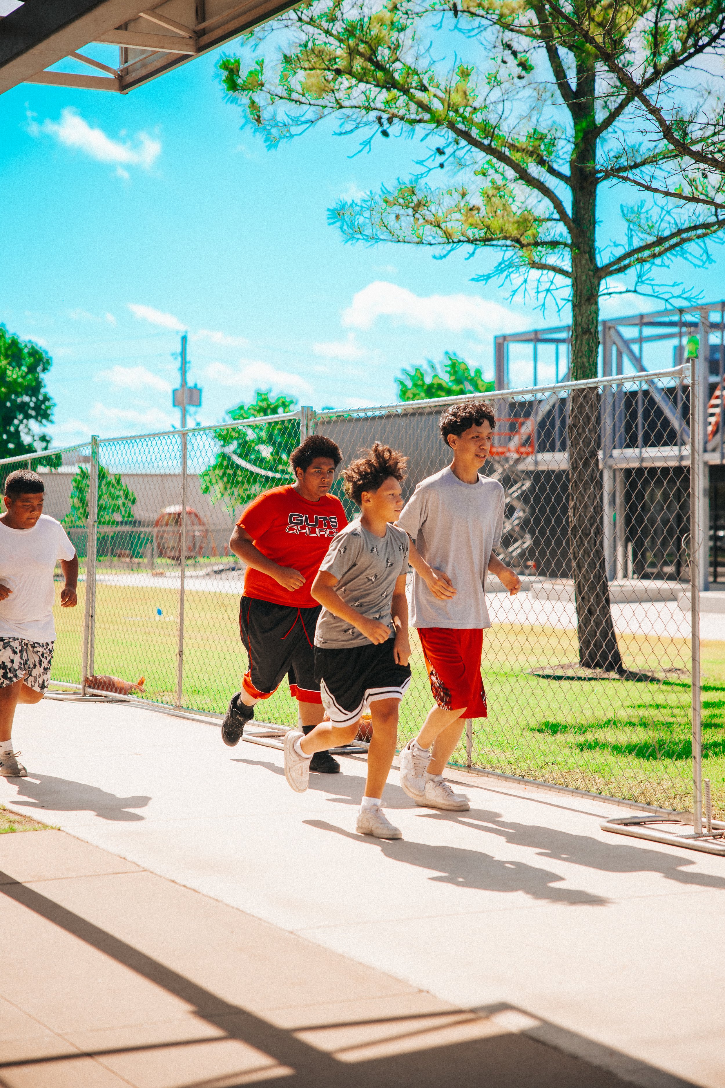 Four young boys running outdoors on a sunny day, with a fence, trees, and a blue sky in the background.