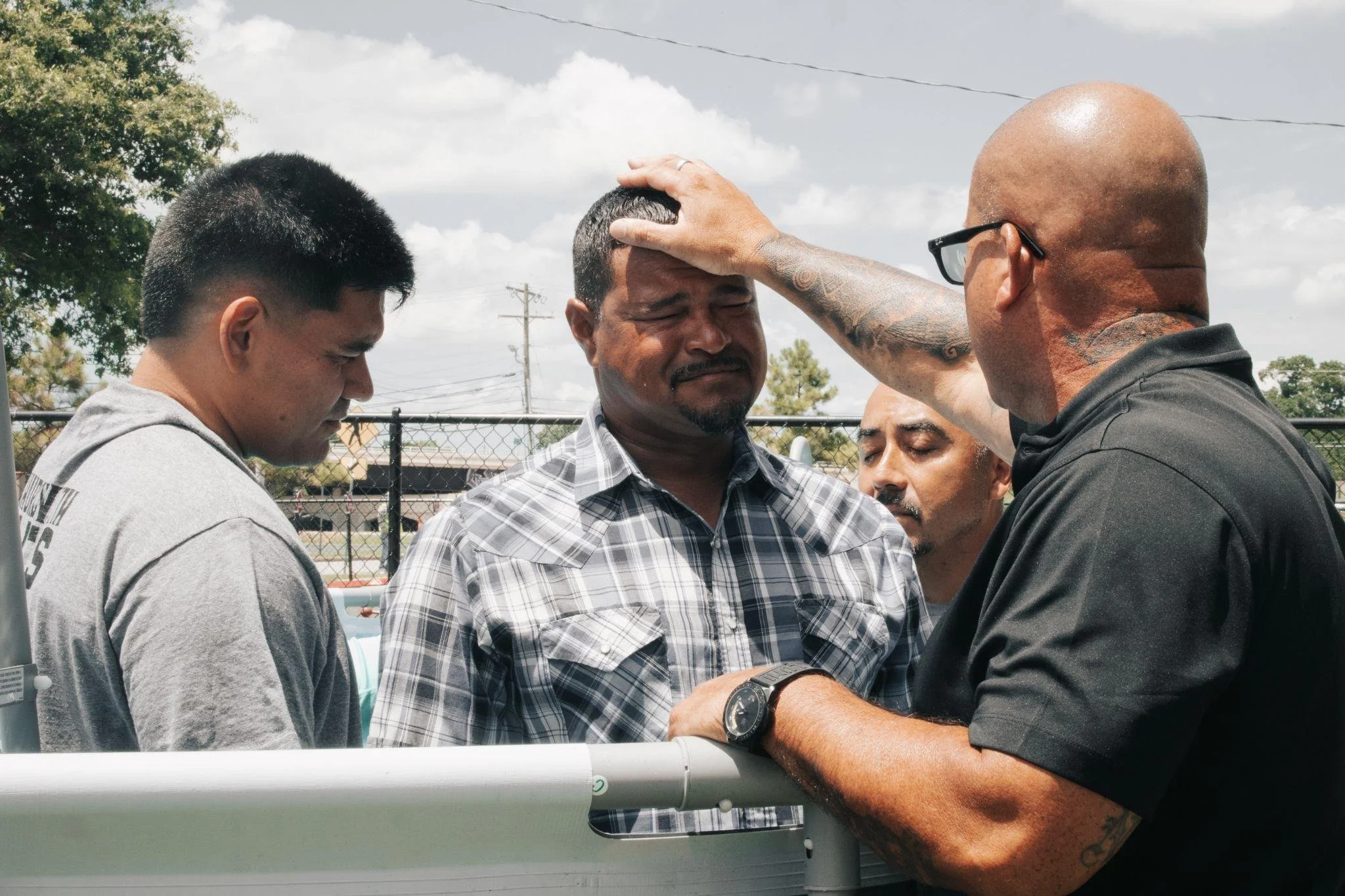 A man with glasses and tattoos on his arm is placing his hand on another man's head in a prayer-like gesture during an outdoor gathering near a fence, with three other men observing and a cloudy sky in the background.