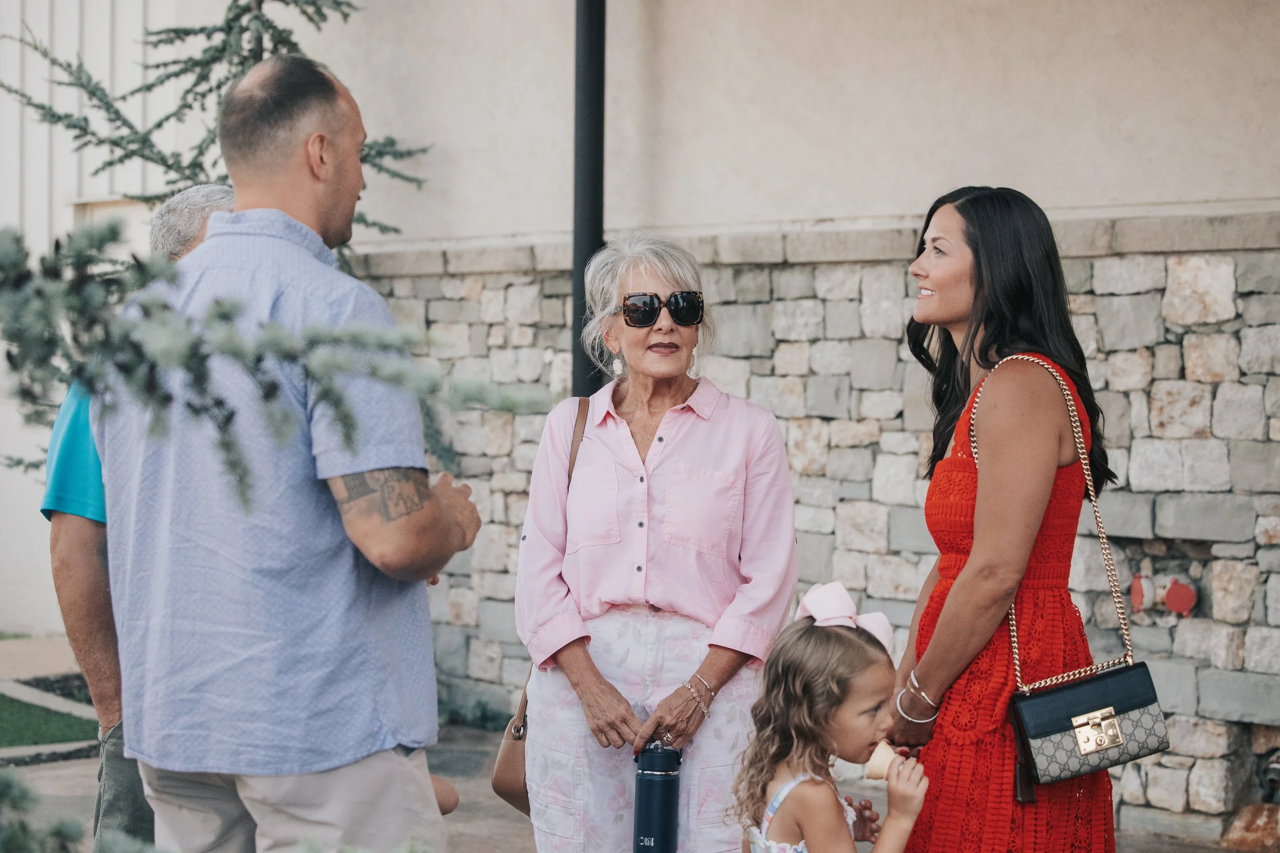 A group of people, including a woman in a red dress, an elderly woman in pink with sunglasses, and a young girl eating ice cream, are talking outdoors in front of a stone wall.
