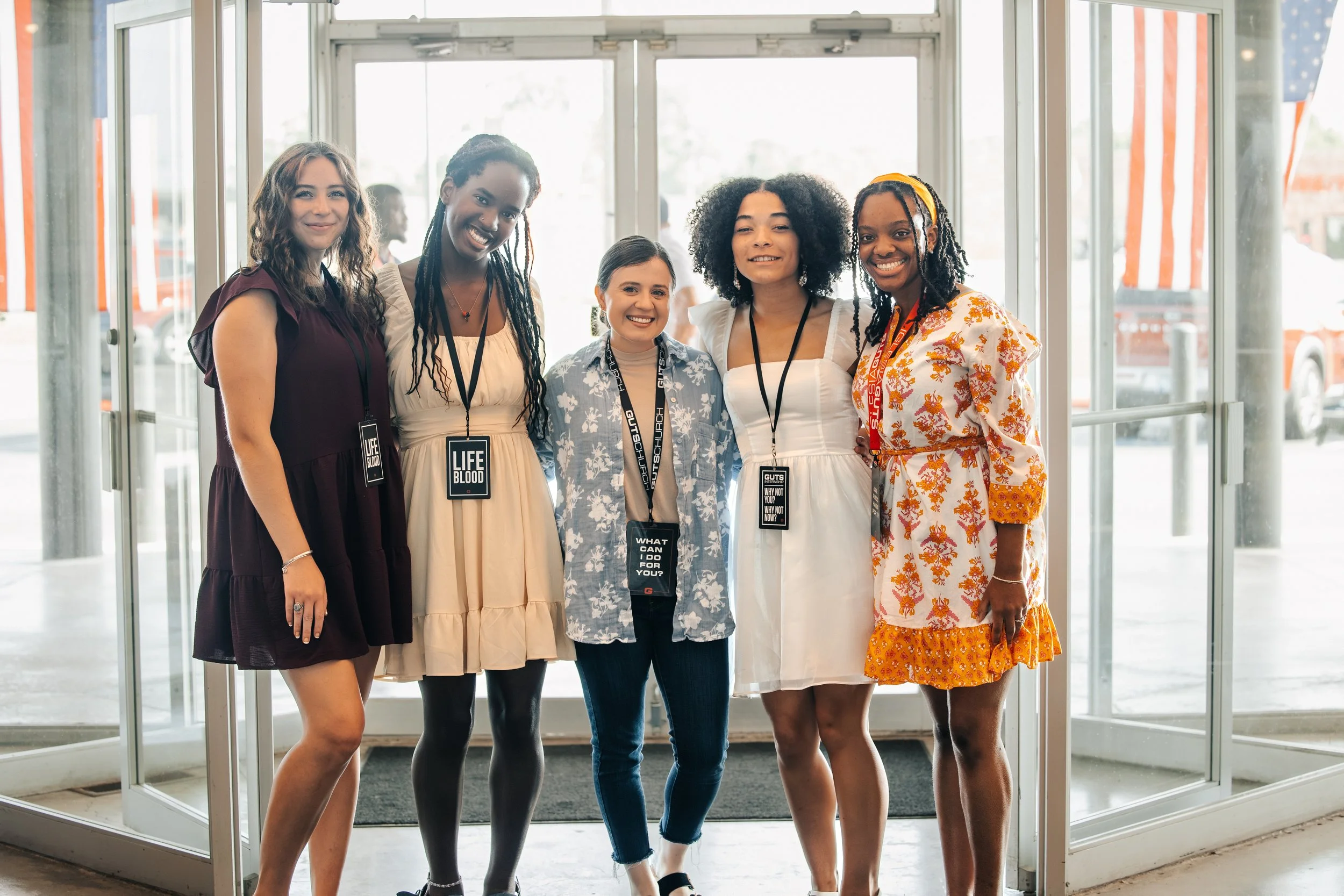 Group of five women standing together in front of a glass entrance door, smiling for the photo, with lanyards and badges around their necks.