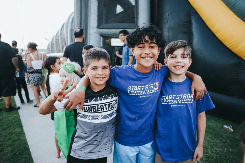 Three boys smiling and hugging at an outdoor event, with a crowd in the background and an inflatable structure to the side.