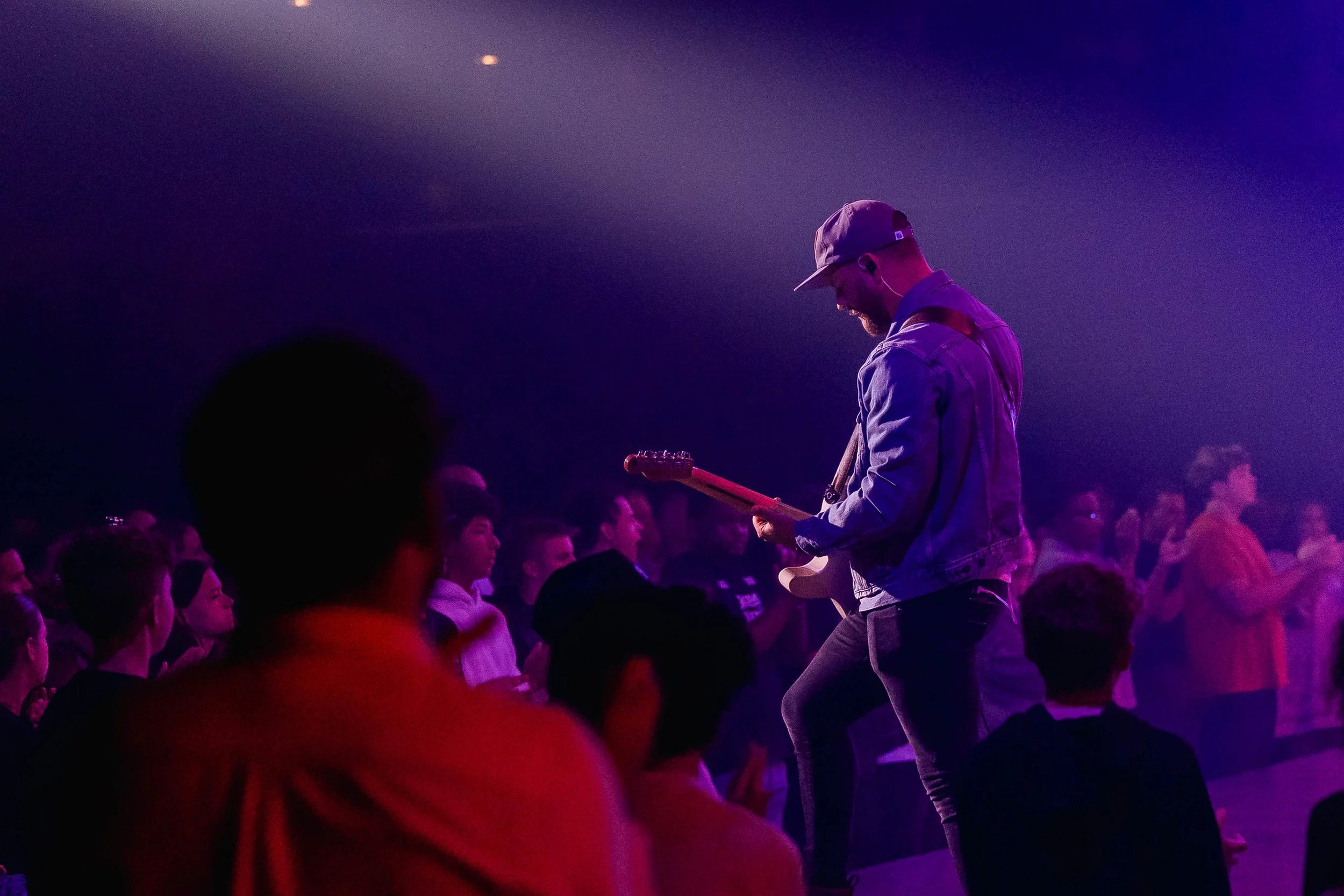 A musician playing an electric guitar on stage in front of a crowd at a concert, illuminated by purple and blue stage lights.