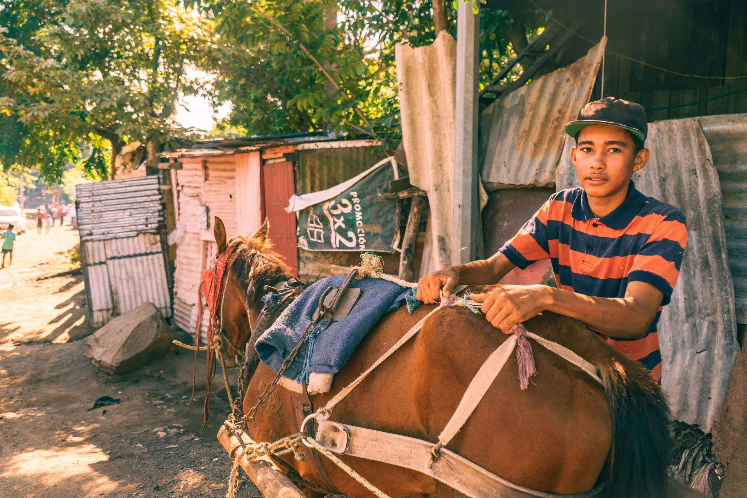 A young man with a striped polo shirt and a cap tending to a horse in a rustic outdoor setting with makeshift structures and trees.