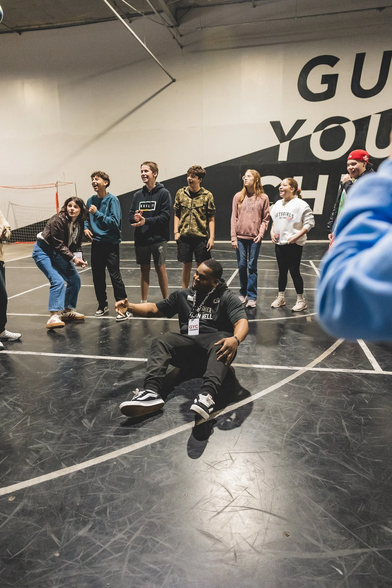 Group of young people and an adult man in a gymnasium, some standing and one sitting on the floor, with a large wall saying 'GUN' and 'YOU' partially visible.