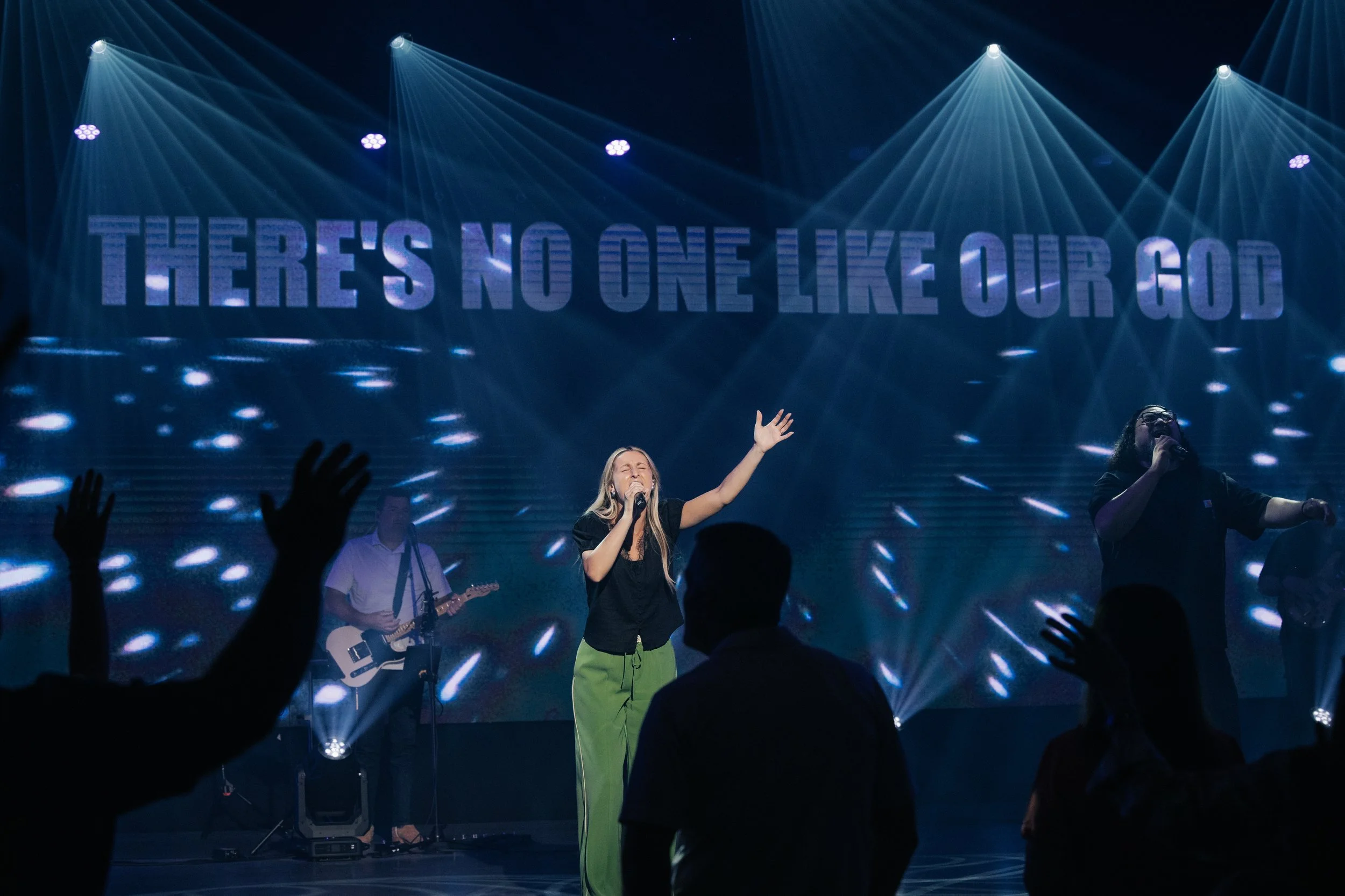 A woman and man singing on stage with hands raised, with a large illuminated sign behind them that says 'THERE'S NO ONE LIKE OUR GOD.'