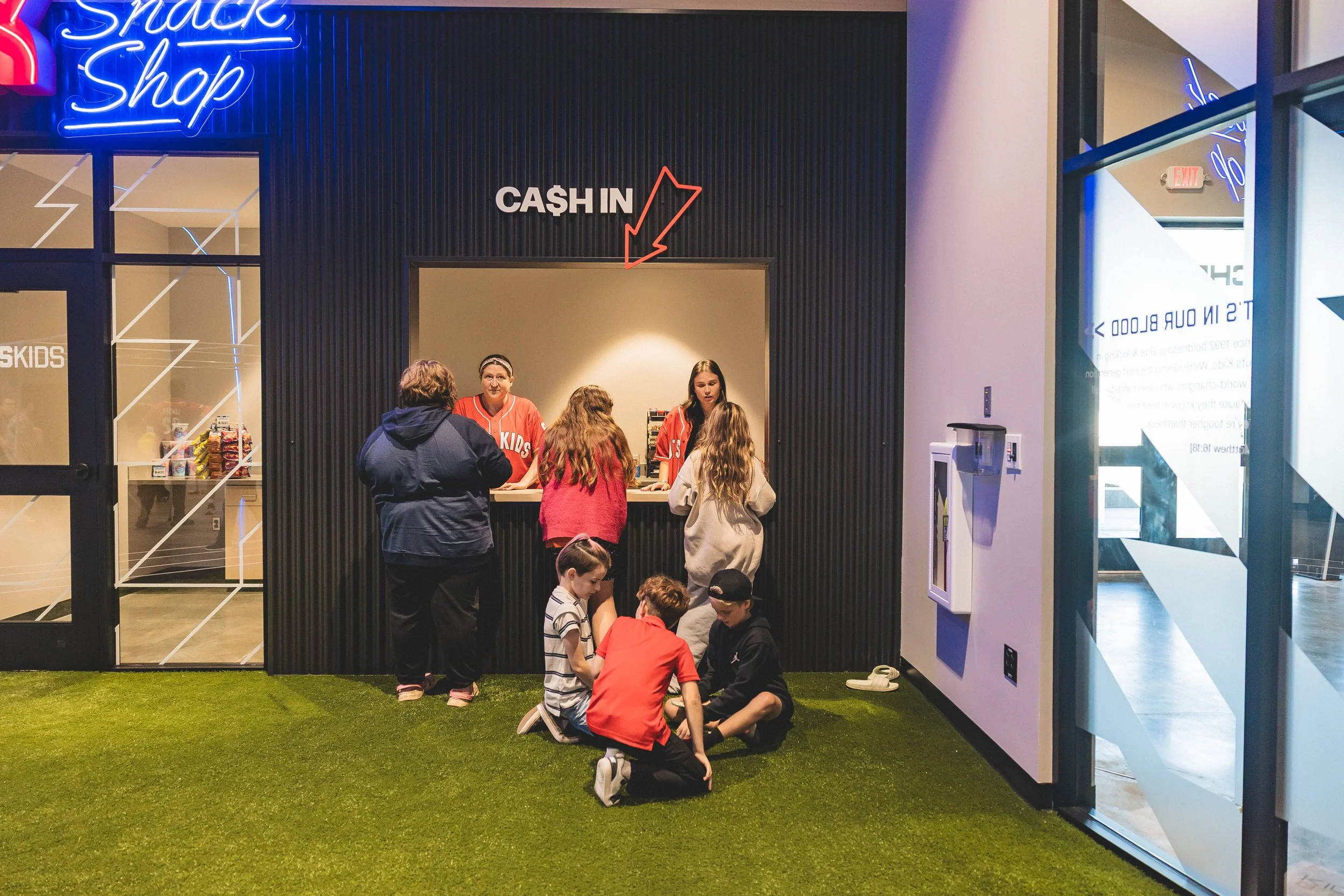 Kids snack shop counter with two kids ordering and three staff members behind the counter, sign above reads 'Cash In' and neon sign says 'Snack Shop'.
