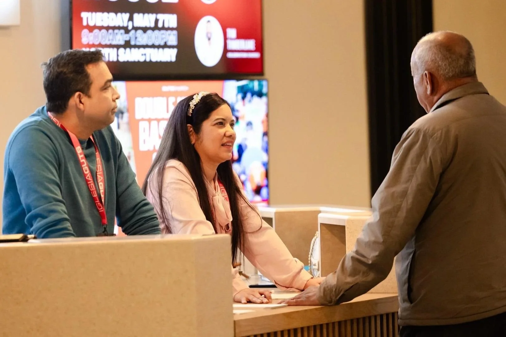 Three people at a reception desk conversing. A young man and woman sit behind the counter, and an older man stands in front of them. Background displays an electronic sign with event details.