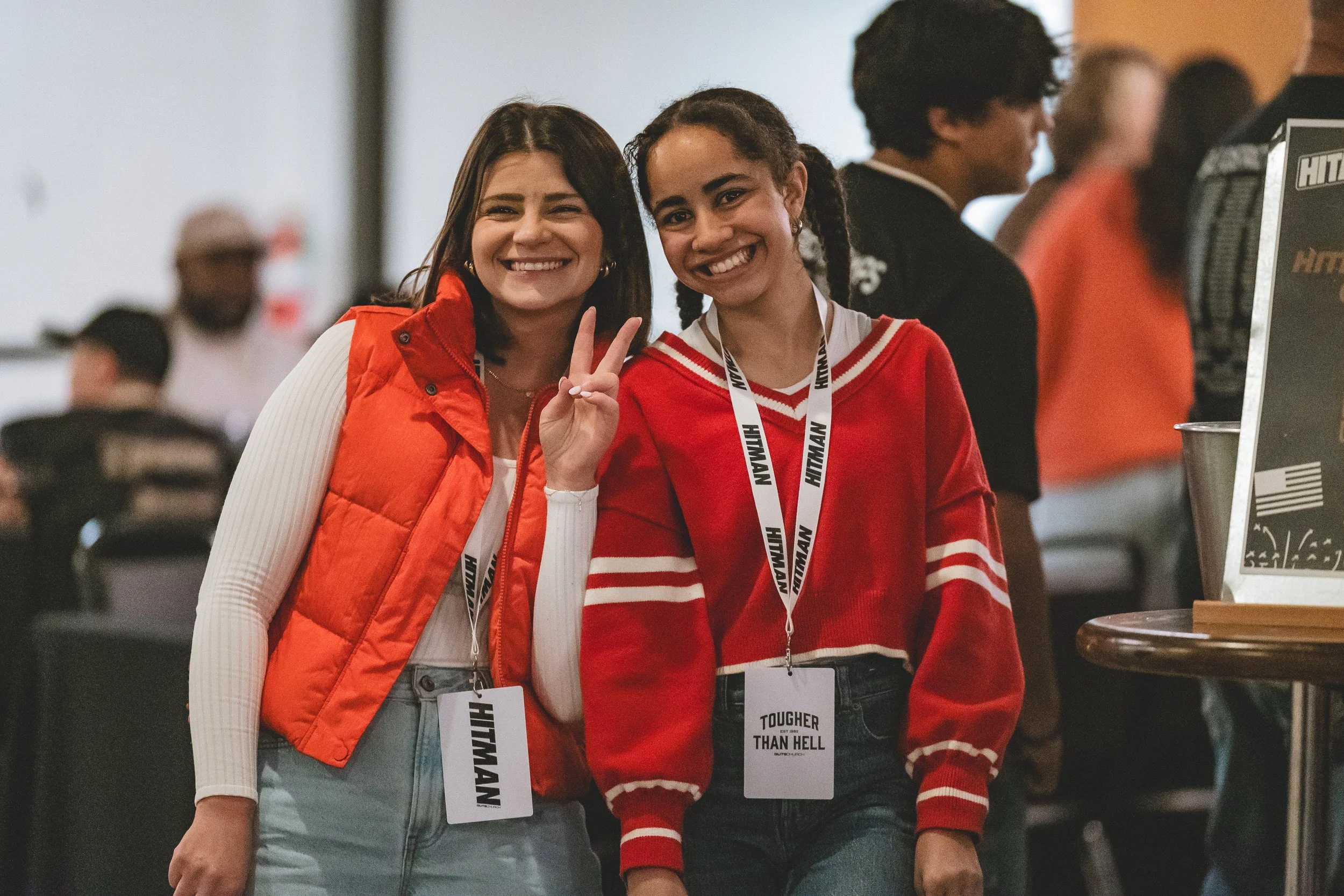 Two young women smiling and making peace signs at a crowded event. One wears a red vest over a white top, the other in a red and white striped sweater. They have event badges hanging around their necks.