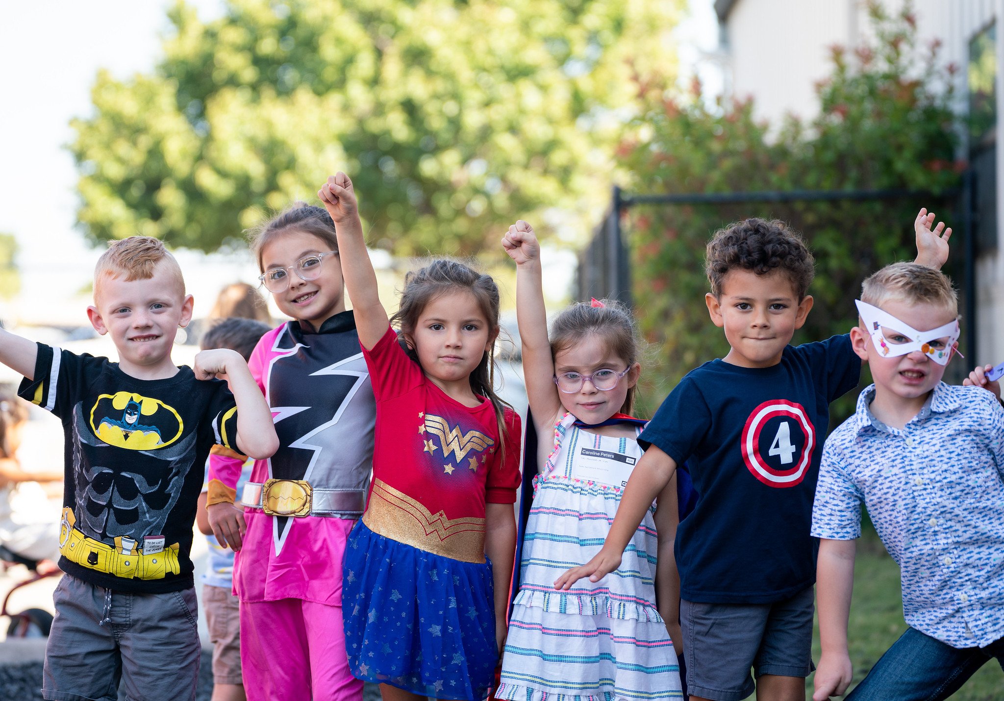 Group of six children dressed in superhero and costume outfits, raising their fists and smiling outdoors in a park.