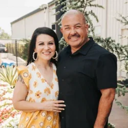 A woman and a man standing outdoors, smiling and posing for the photo, with a garden and a building in the background.