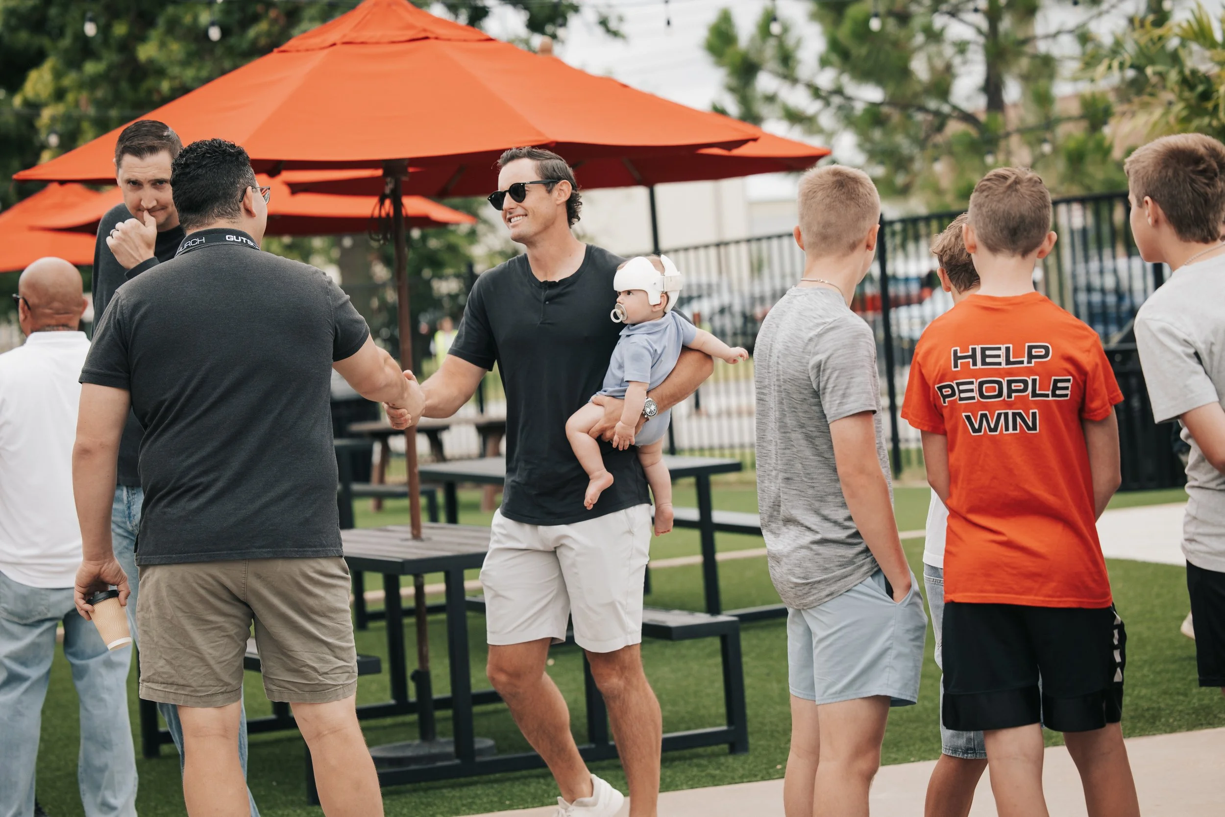 People shaking hands outdoors near orange umbrellas and picnic tables with a group of children nearby.
