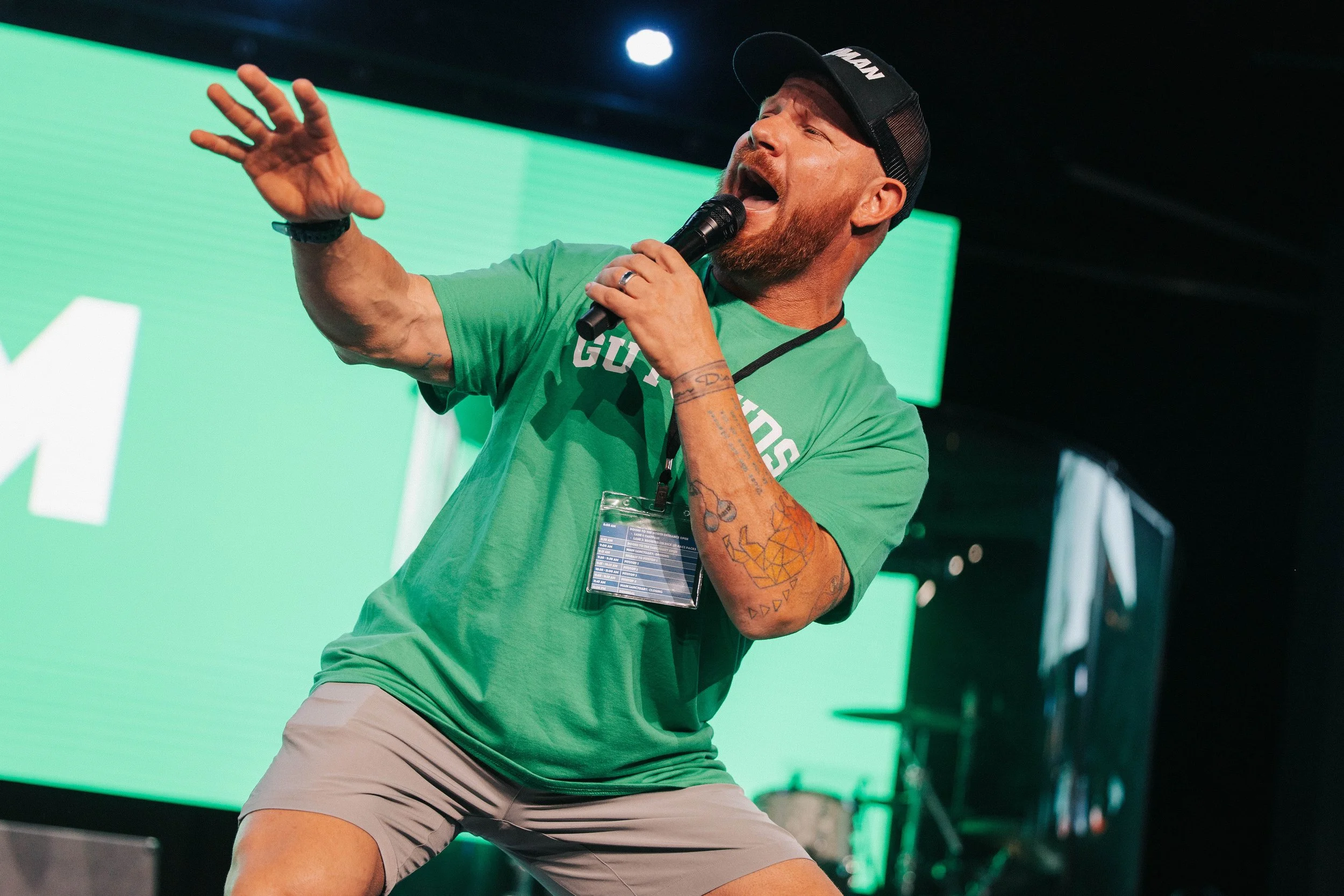A man with a beard and tattoos on his left arm is singing into a microphone on stage. He is wearing a green T-shirt, khaki shorts, and a black baseball cap. There is a large green screen behind him and some stage equipment visible.