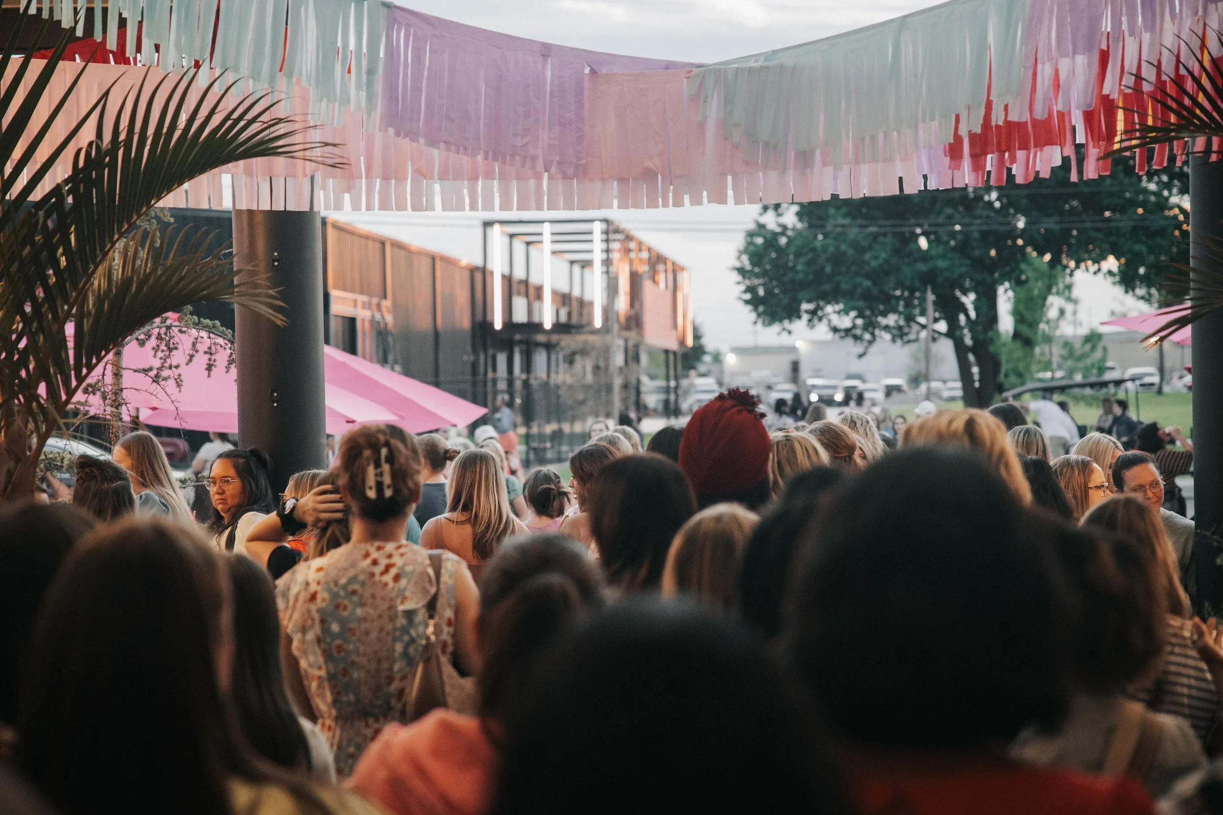 Crowd of people gathered outdoors under pastel-colored streamers and pink umbrellas, with trees and modern buildings in the background.