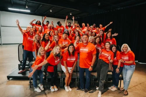Group of diverse people wearing matching orange shirts, smiling and posing together indoors, some with their arms raised.
